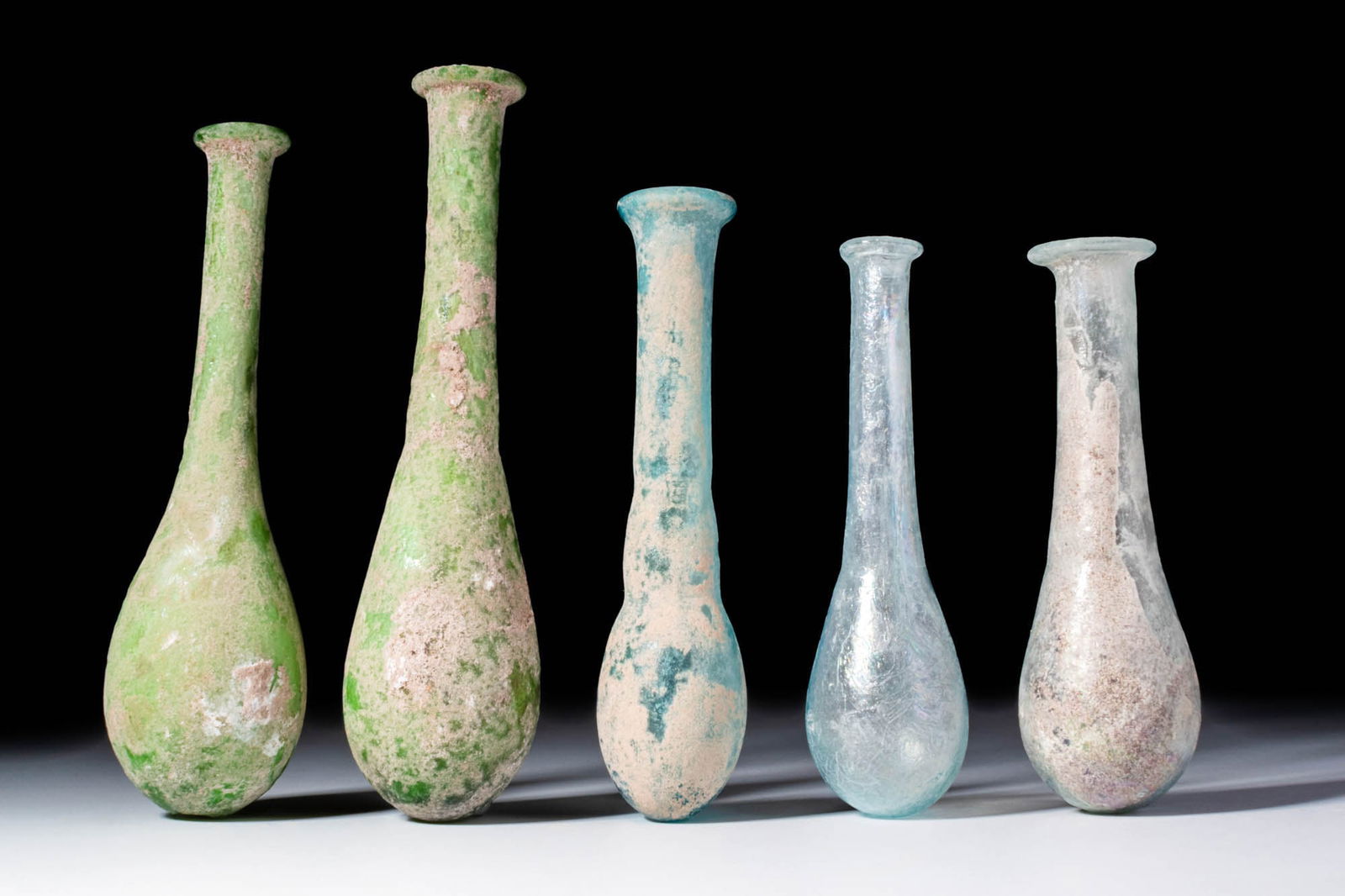 COLLECTION OF ROMAN GLASS UNGUENTARIA: Ca. AD 1 - 300. A collection of five blown glass unguentaria, each with pear-shaped lower body and elongated cylindrical neck leading to a pronounced, rounded rim. The glass varies from translucent pa