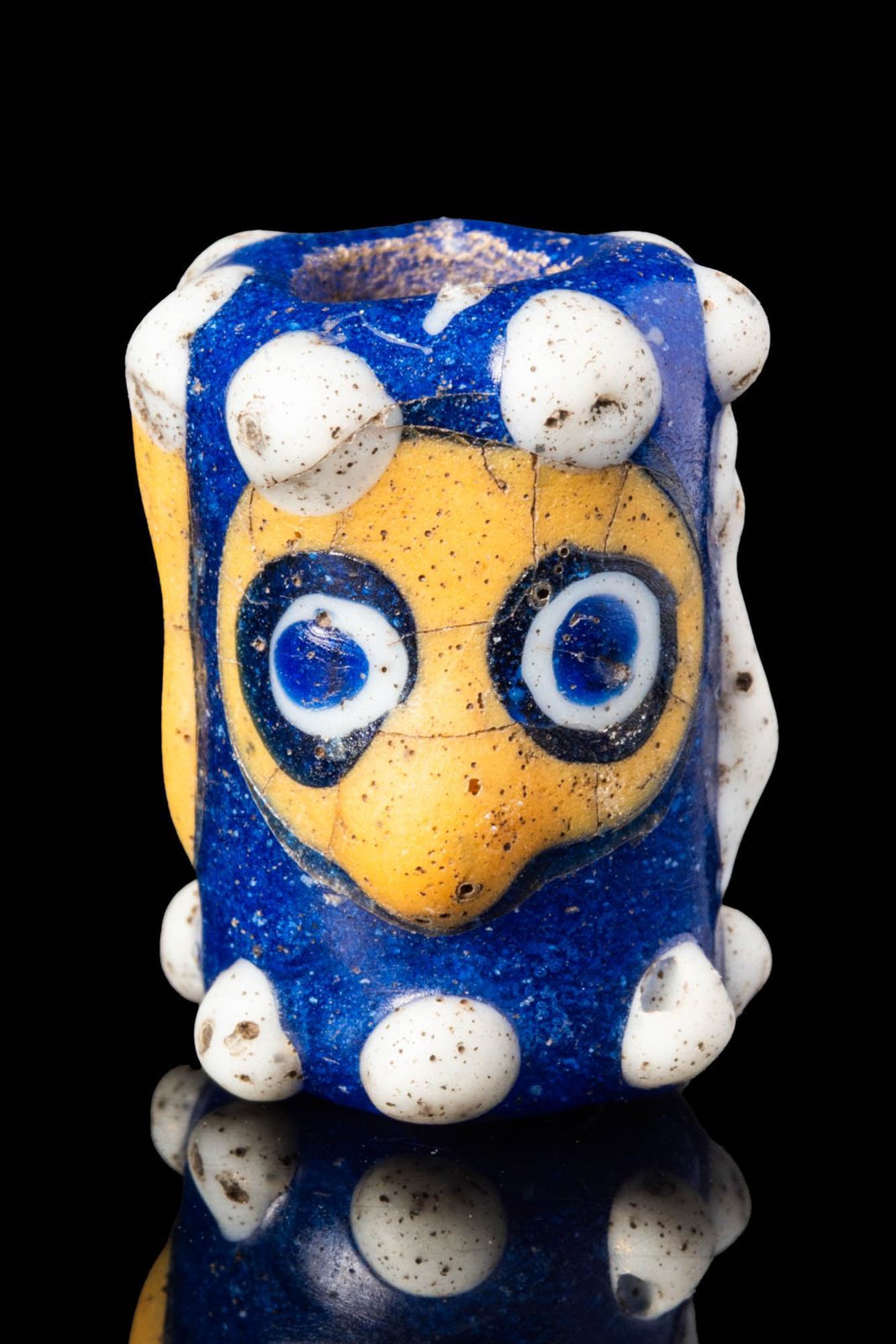 HUGE PHOENICIAN POLYCHROME GLASS HEAD-SHAPED BEAD: Ca. 600 - 400 BC. A brightly coloured mosaic glass tubular bead with stylised faces on opposing sides: one rendered in opaque yellow glass, the other in white. Each face features large, circular cobal