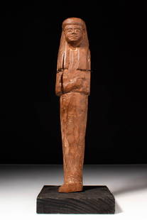 EGYPTIAN WOODEN USHABTI RAMESSIDE PERIOD: New Kingdom, Ca. 1292 - 1189 BC. A wooden ushabti with a tall, upright form and angular limbs. The face is broad, with incised details defining the eyes, nose, and mouth. The figure wears a tripartite
