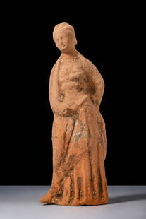 GREEK TANAGRA FEMALE FIGURINE: Ca. 400 - 200 BC. A terracotta statuette of a standing female figure draped in a himation over a chiton, the weight resting on the right leg, the left bent slightly at the knee. The folds of the garme