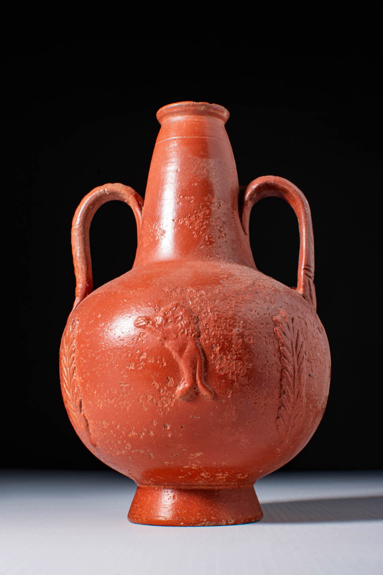 NORTH AFRICAN TERRA SIGILLATA FLASK DECORATED WITH LIONS: Ca. AD 300. A terra sigillata rounded flask with a tall, conical neck and twin loop handles. The body is decorated in relief with lions interspersed with vertical leafy branches. The piece rests on a