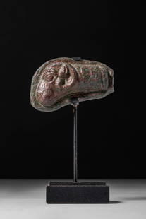 ROMAN BRONZE PLAQUETTE WITH RAM’S HEAD: Ca. AD 1 - 300. A bronze fitting in the form of a ram’s head, shown in profile with a prominent curved horn, circular eye, and rounded muzzle. The back of the fitting is flat. Mounted on a modern st