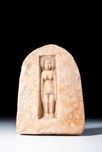 PHOENICIAN VOTIVE STELE WITH GODDESS TANIT: Ca. 600 - 500 BC. A limestone votive stele with a rounded top, depicting a standing female figure carved in shallow relief within a rectangular niche. The figure is shown nude, with hands along the bo