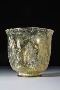ROMAN GREEN GLASS BEAKER: Ca. AD 100 - 300. A translucent green glass beaker with a rounded body tapering slightly towards a small foot. The walls are decorated with shallow vertical indentations, and the rim is uneven and
