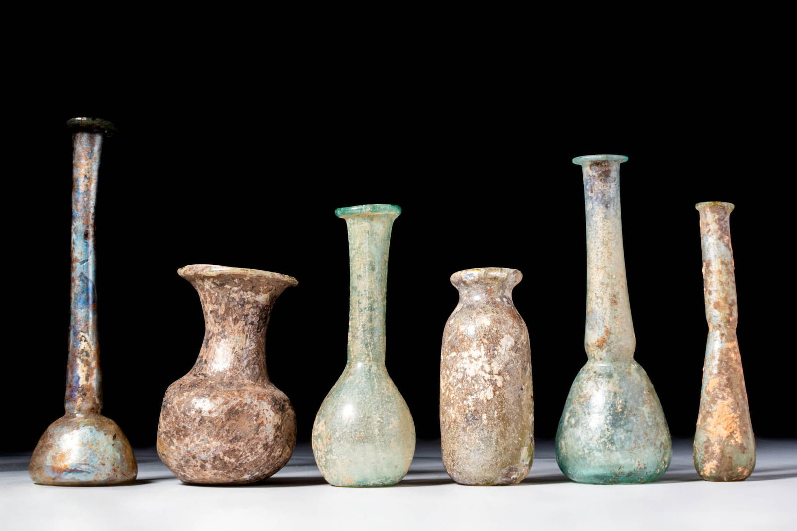 COLLECTION OF ROMAN GLASS UNGUENTARIA: Ca. AD 1 - 300. A collection of six glass unguentaria. The first on the left has a long neck and domed lower body, covered in a bluish iridescent crust. The second has a funnel-shaped neck and a sprea