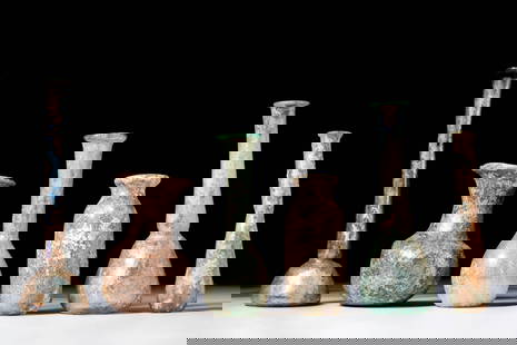 COLLECTION OF ROMAN GLASS UNGUENTARIA: Ca. AD 1 - 300. A collection of six glass unguentaria. The first on the left has a long neck and domed lower body, covered in a bluish iridescent crust. The second has a funnel-shaped neck and a