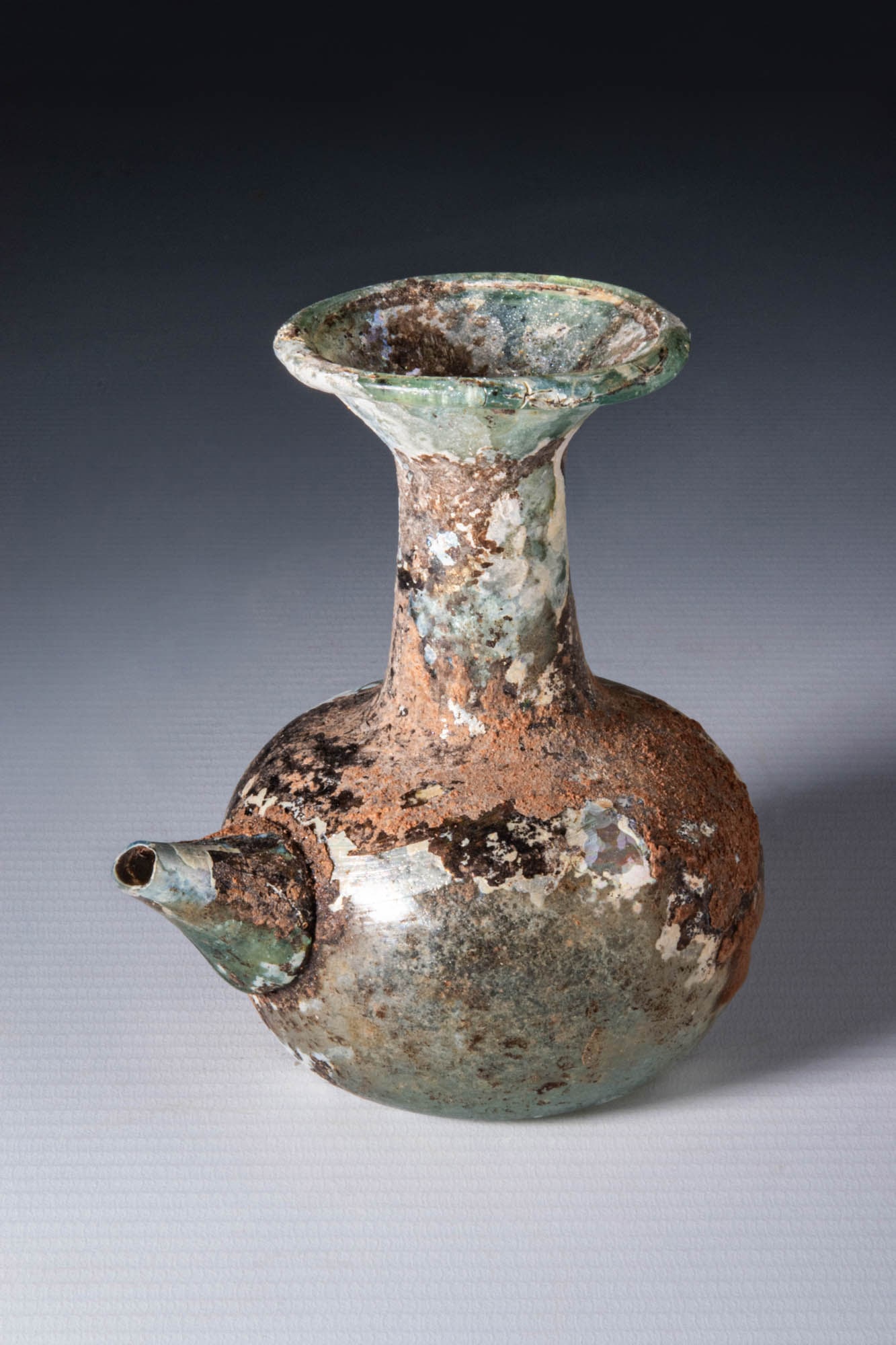 ROMAN GLASS INFUNDIBULUM (LAMP FILLER): Ca. AD 100 - 200. A free-blown glass flask with an apple-shaped lower body and a long, cylindrical neck that ends in a flared mouth with a thick, pronounced rim. The base is flattened with a slight ce