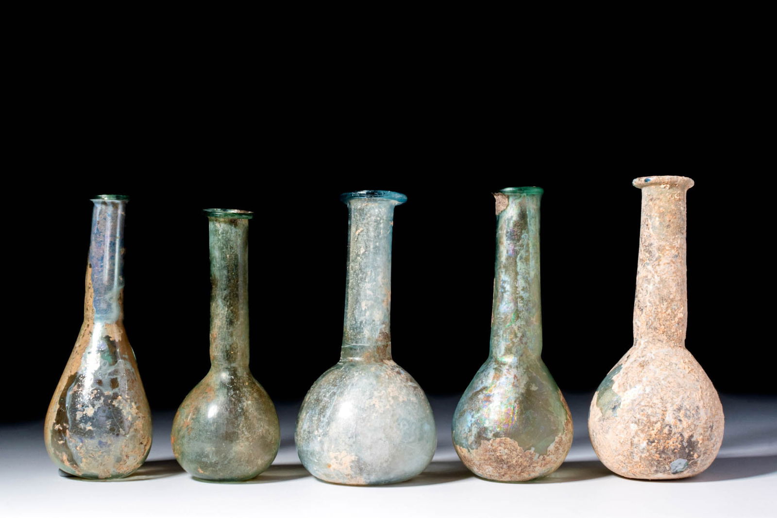COLLECTION OF ROMAN UNGUENTARIA: Ca. AD 1 - 300. A collection of five blown glass unguentaria, each with a pear-shaped lower body and elongated cylindrical neck leading to a pronounced, rounded rim. The glass varies from translucent