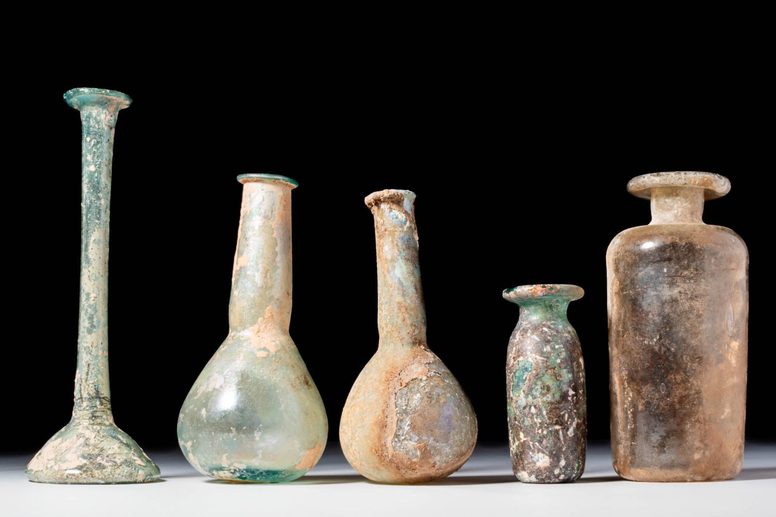 COLLECTION OF ROMAN GLASS UNGUENTARIA AND BOTTLE: Ca. AD 1 - 300. A collection of three glass unguentaria and two bottles. The first on the left has a tall, cylindrical neck and a spreading foot. The second and the third have pear-shaped bodies with