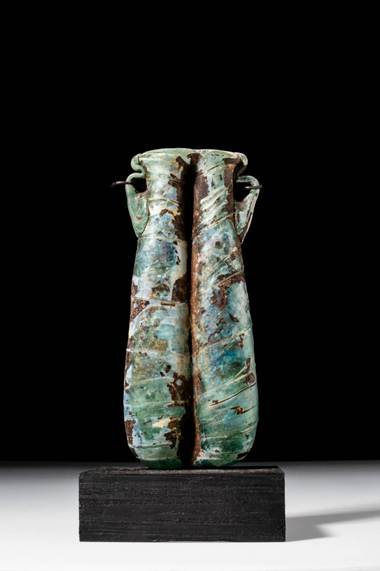 ROMAN GLASS DOUBLE UNGUENTARIUM: Ca. AD 300 - 500. A translucent green glass double unguentarium formed from two elongated cylindrical tubes joined along their lengths. A single applied trail spirals around both. Each vessel flares s