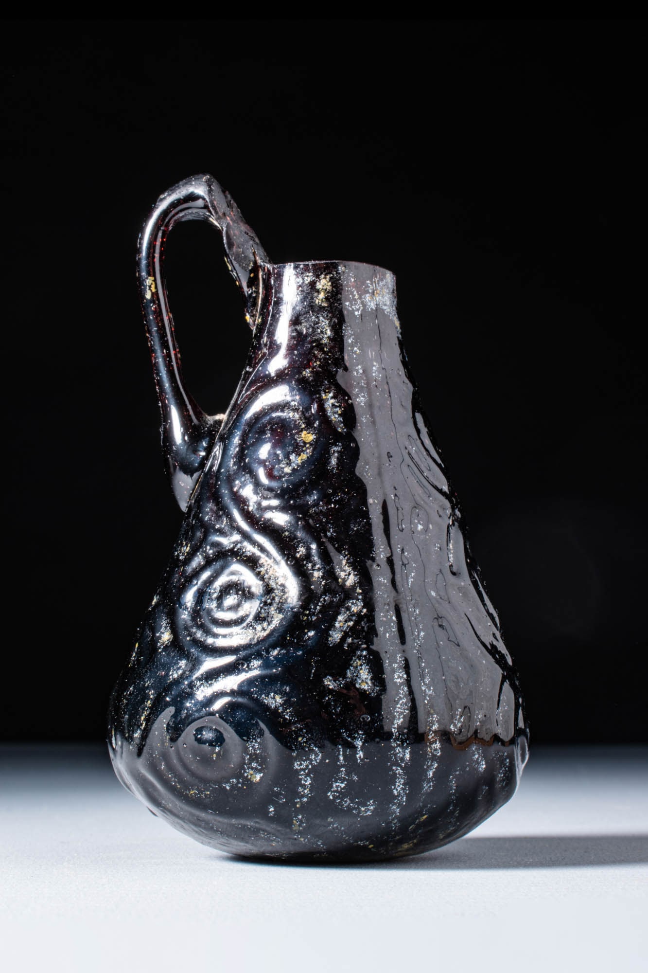 LATE ROMAN AUBERGINE GLASS JUG WITH MOULD-BLOWN DECORATION: Ca. AD 300 - 500. A glass jug with a pear-shaped body decorated with mould-blown raised circular and linear motifs. The body tapers towards the opening, and a single applied handle extends from the sh