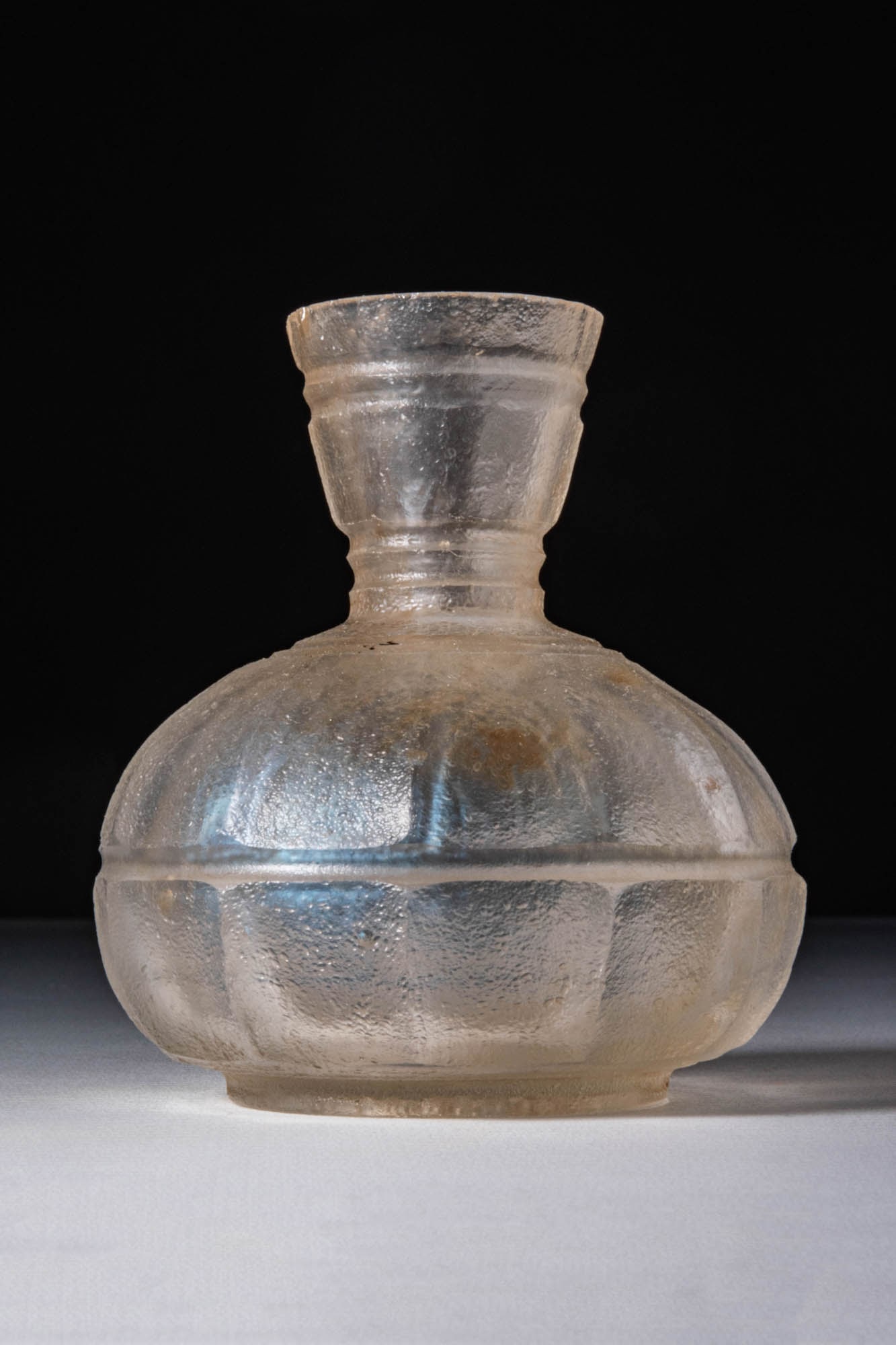 SASANIAN OR EARLY ISLAMIC GLASS UNGUENTARIUM: Ca. AD 600 - 800 . A translucent flask with a broad, rounded body segmented by vertical ribs and a ring base. The narrow neck is marked by two shallow grooves and expands into a flaring, funnel-shaped