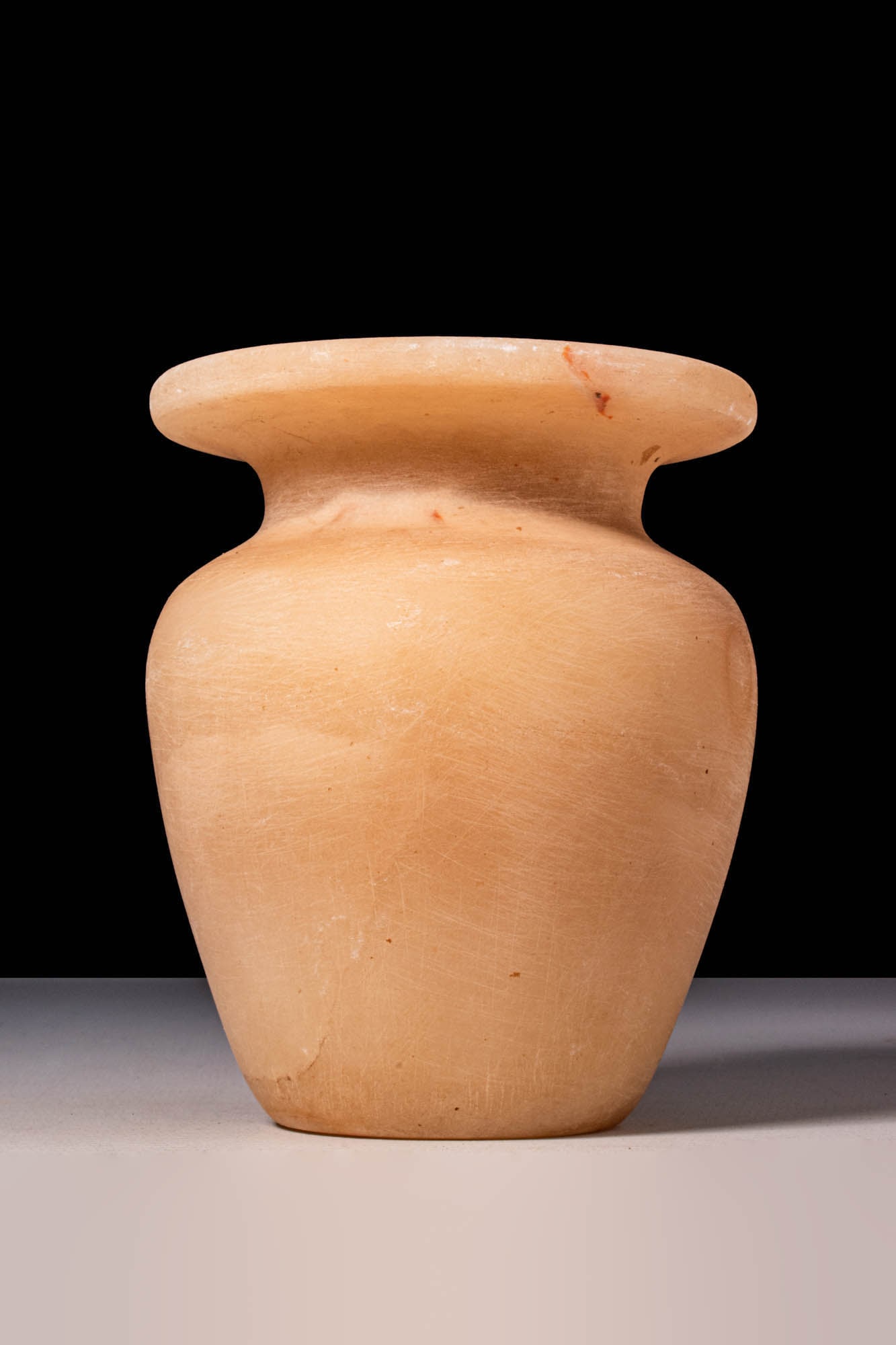 EGYPTIAN ALABASTER VOTIVE VESSEL: New Kingdom, Ca. 1550 - 1069 BC. A vessel carved from alabaster with a rounded body tapering to a narrow base. The shoulders curve gently inward to a short neck, surmounted by a wide, flat rim with an