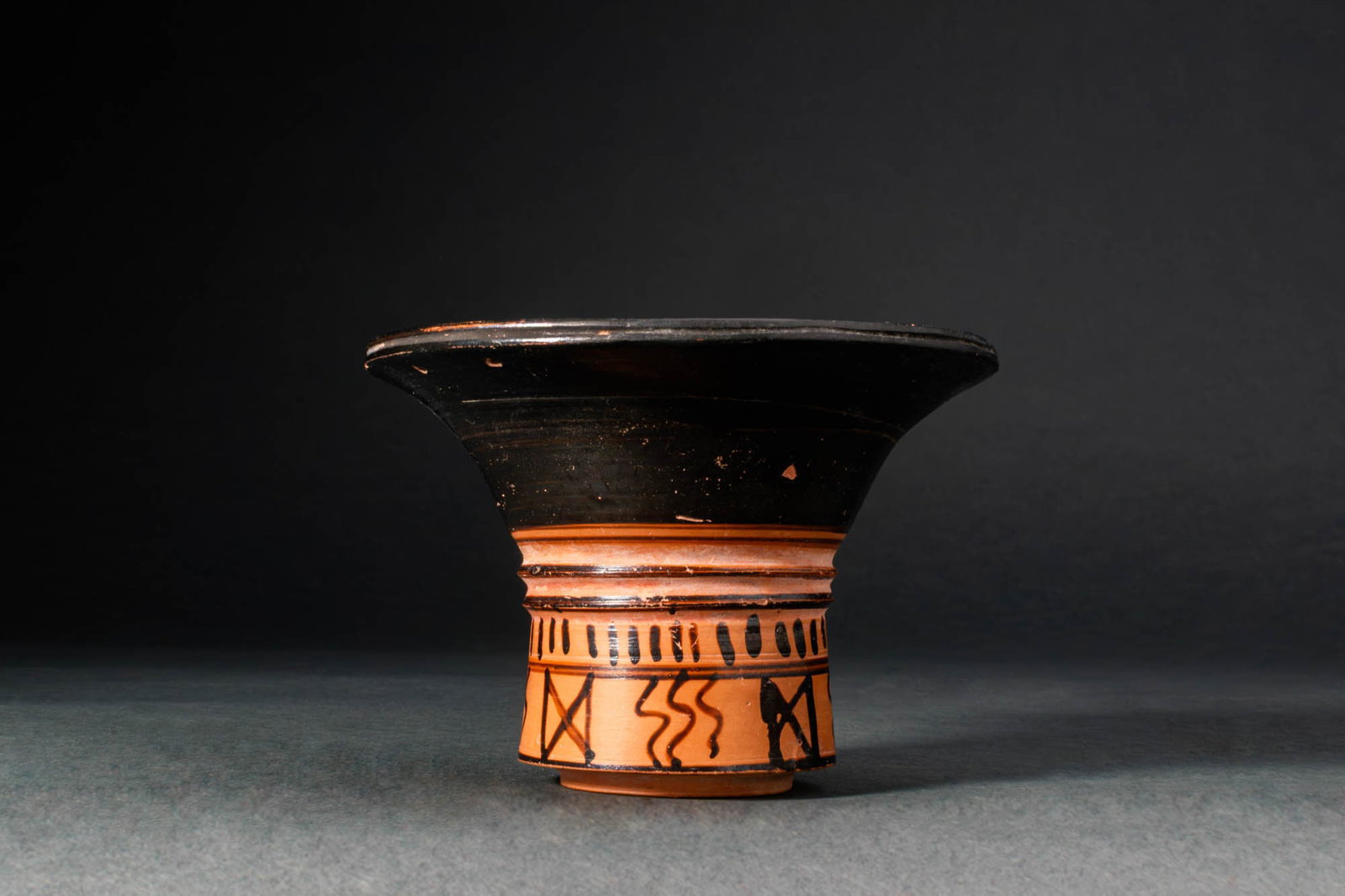 RARE GREEK KALATHOS WITH GEOMETRIC MOTIF: Ca. 600 BC. A terracotta kalathos with a sharply flaring rim, the interior and exterior coated in a lustrous black glaze, set above a short cylindrical body left unglazed. The midsection is encircled 