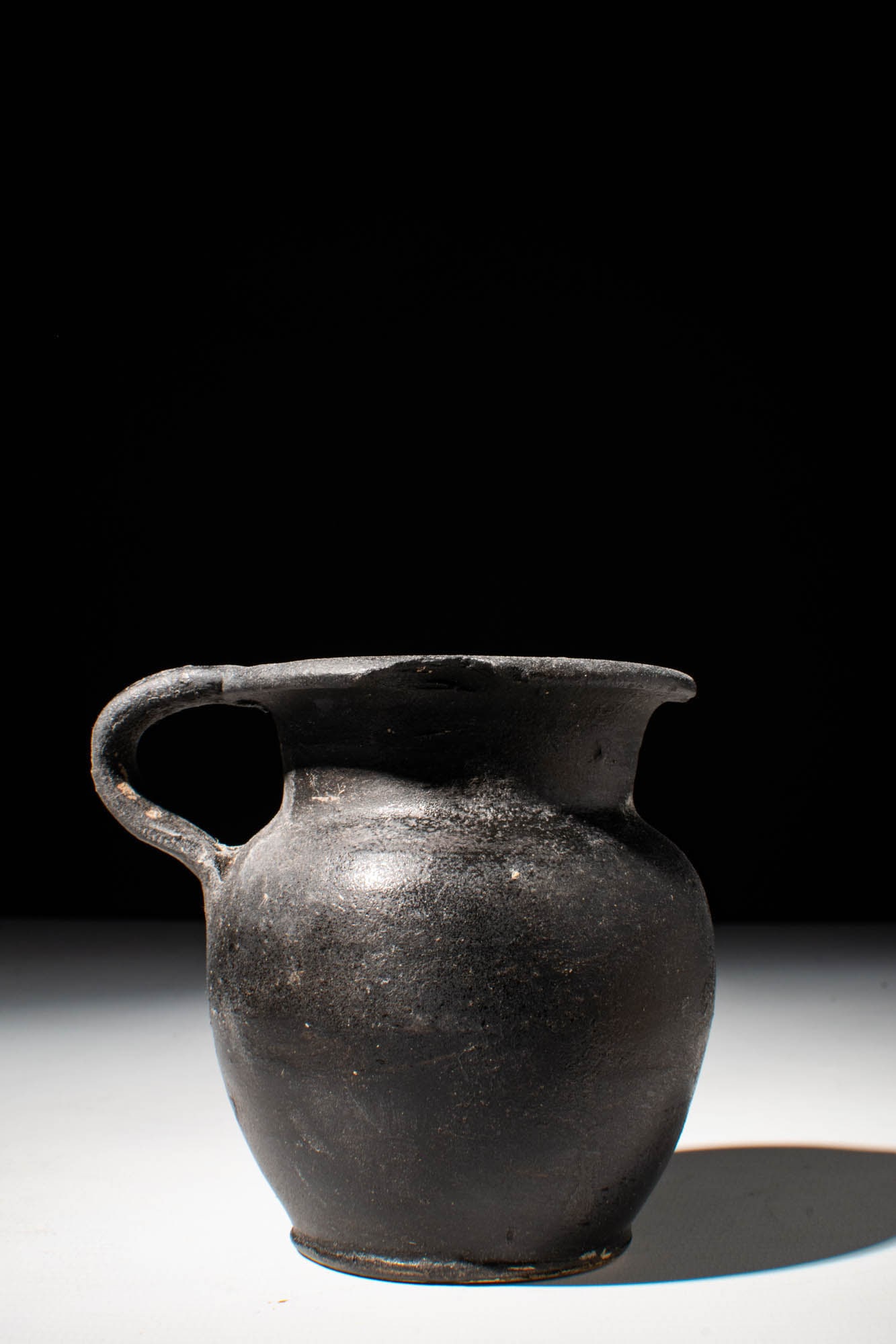 GREEK BLACK GLAZE JUG: Ca. 400 - 300 BC. A black glaze terracotta jug. The piece is of good size and has an ovoid body, a short wide neck with a flaring rim, featuring a wide strap handle from rim to shoulder and a flat bas