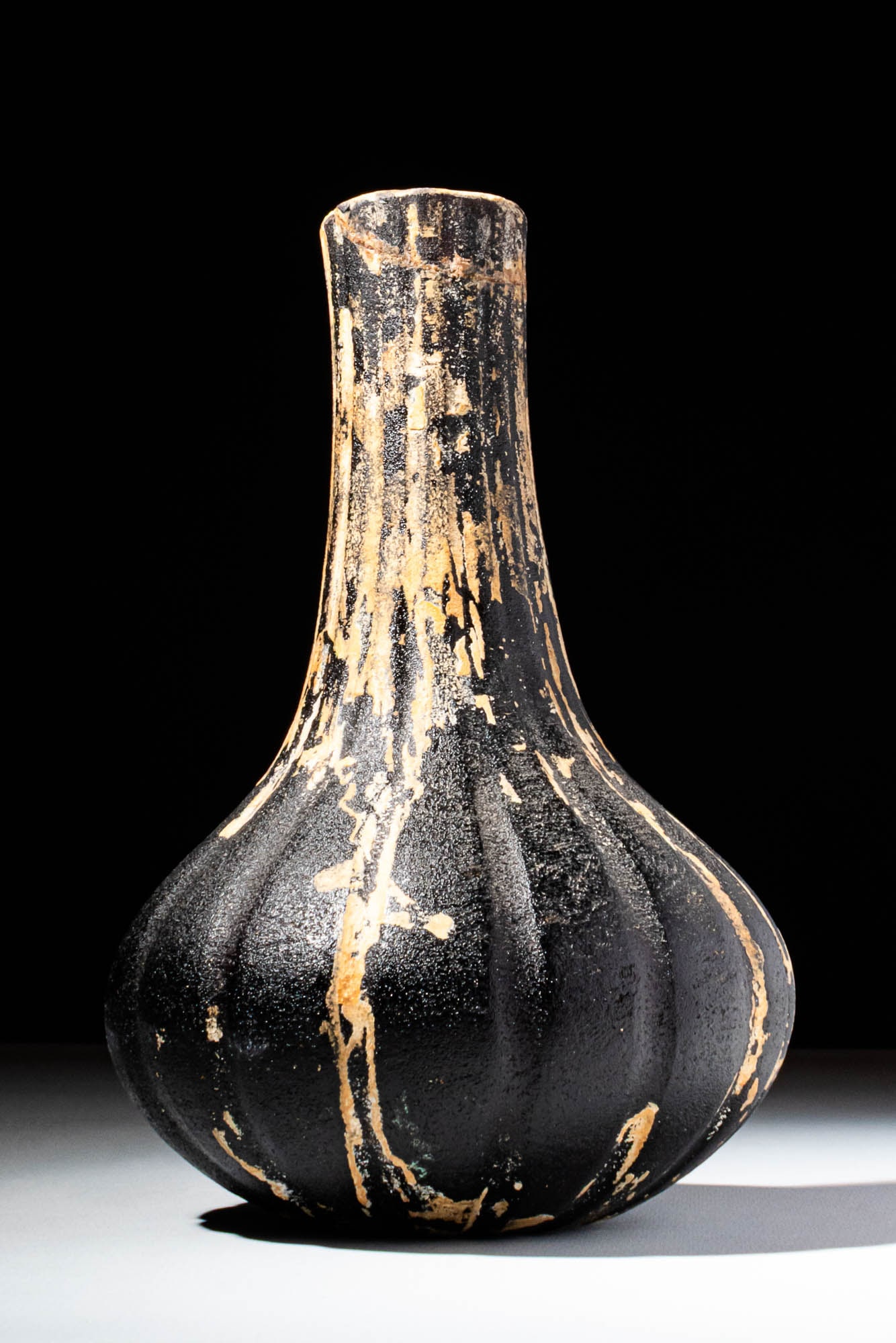 ROMAN AUBERGINE GLASS RIBBED FLASK: Ca. 100 BC - AD 100. An aubergine-coloured glass bottle with an onion-shaped body, elongated cylindrical neck, and plain, upright rim. The body is subtly faceted with vertical ribbing that converges o