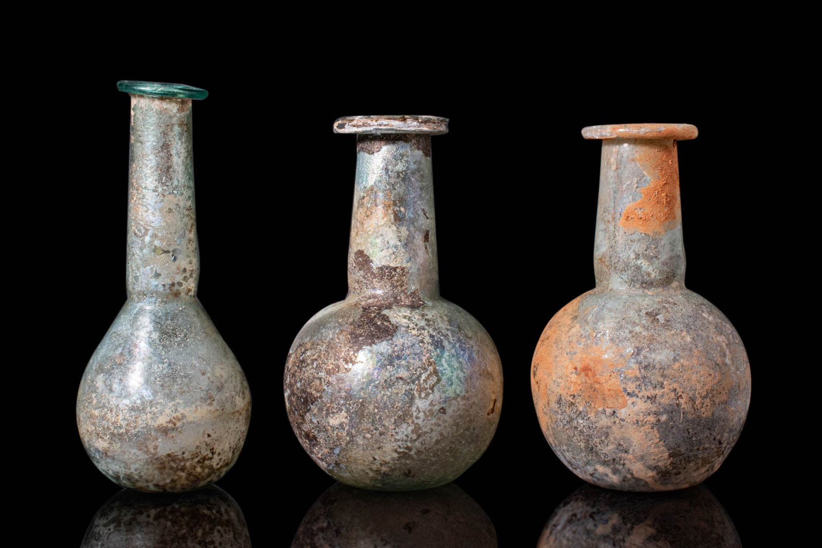 COLLECTION OF ROMAN GLASS UNGUENTARIA: Ca. AD 200 - 400. A collection of three glass unguentaria. The leftmost vessel has a piriform body, a tall neck, and a rounded rim. The other two pieces share a compact form with squat necks, evenly r