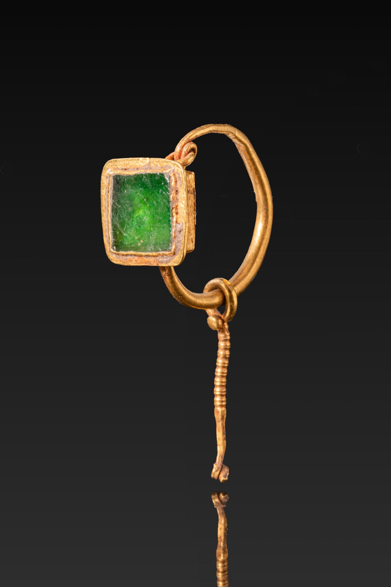 BYZANTINE GOLD EARRING WITH SQUARE BEZEL: Ca. AD 400 - 600. A gold earring formed of a circular hoop attached to a rectangular box-shaped bezel set with a flat, square-cut green gem framed by a narrow rim. A coiled wire rod is suspended from