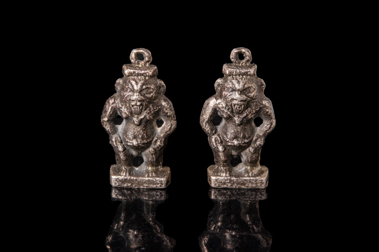 PAIR OF EGYPTIAN SILVER BES AMULETS: Third Intermediate Period, Ca. 1069 - 664 BC. A pair of silver Bes amulets, each modelled as the dwarf_god squatting on an integral rectangular plinth, hands braced on the thighs, and a leonine face w