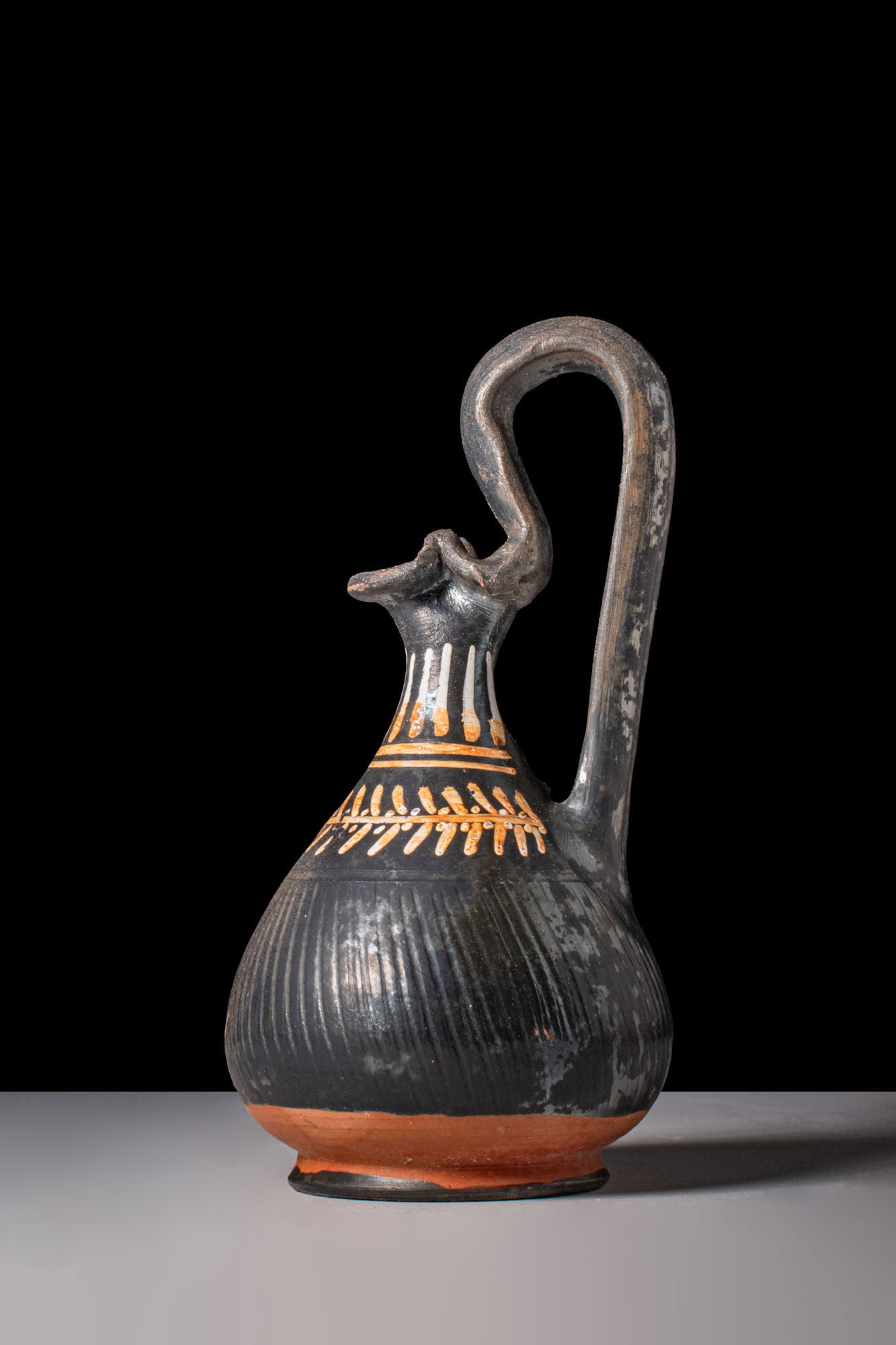 ELEGANT GNATHIAN OINOCHOE WITH LEAVES MOTIF: Ca. 330 - 300 BC. A black-glazed pottery oinochoe with a trefoil mouth above a tall neck and a pear-shaped body on a low ring foot. The lower body is decorated with closely spaced vertical ribbing; th
