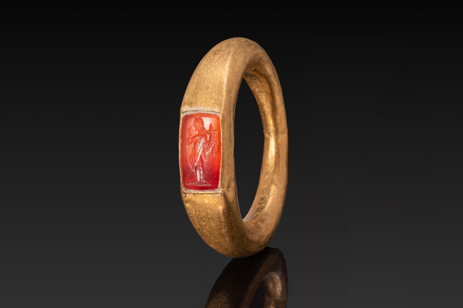 ROMAN CARNELIAN INTAGLIO DEPICTING FORTUNA IN GOLD RING: Ca. AD 50 - 200. A red carnelian intaglio engraved with Fortuna standing left, draped, holding a cornucopia in the left arm. Set within a rectangular recess, in a plain collet sunk into a gold ring of