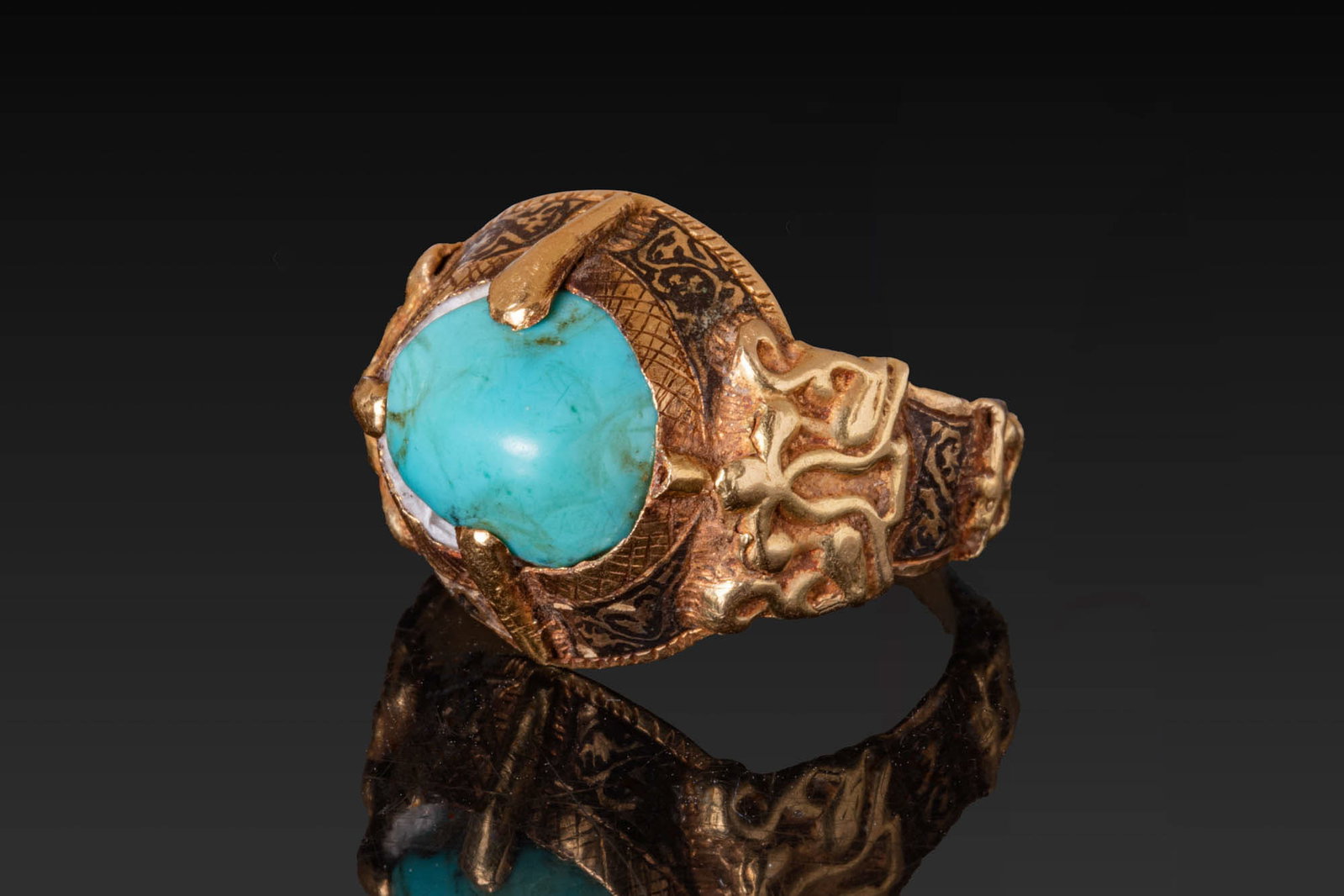 SELJUK GOLD AND NIELLO RING WITH TURQUOISE CABOCHON: Ca. 13th - 15th century AD. A gold and niello ring with a turquoise cabochon secured by four prongs on a high circular box setting encircled by a niello frieze of geometric and foliate motifs. A broad