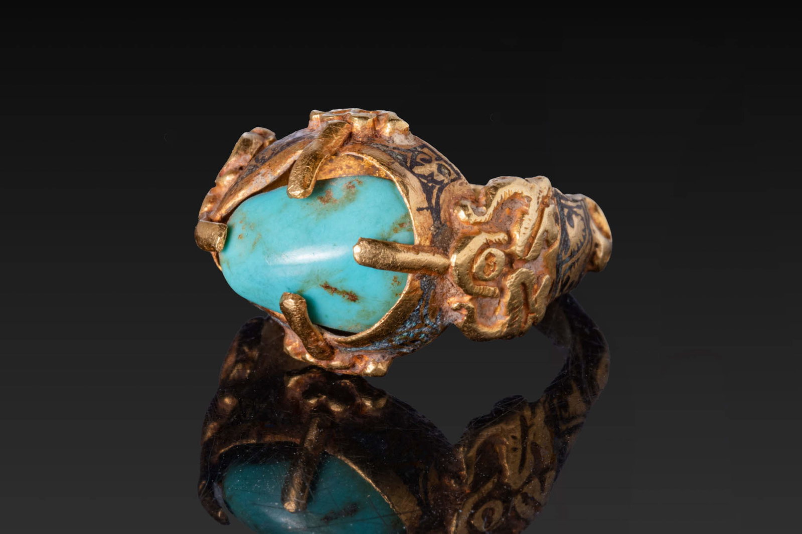 SELJUK GOLD AND NIELLO RING WITH TURQUOISE CABOCHON: Ca. 13th - 15th century AD. A gold and niello finger ring with a broad D-section hoop decorated with raised scrolling ornaments and nielloed panels. It supports an oval box bezel encircled by a niello