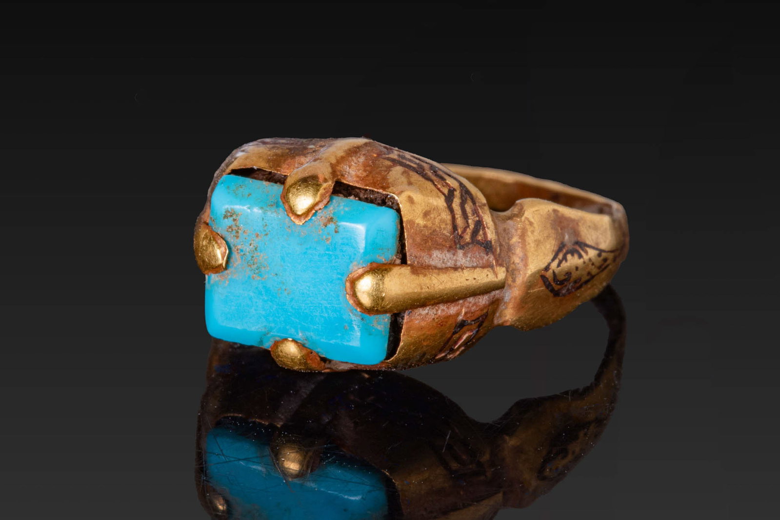SELJUK GOLD AND NIELLO RING WITH TURQUOISE CABOCHON: Ca. 13th - 15th century AD. A gold finger ring with a round hoop that broadens to angular shoulders, each embellished with niello ornament. The high box bezel has four prominent prongs securing a rect