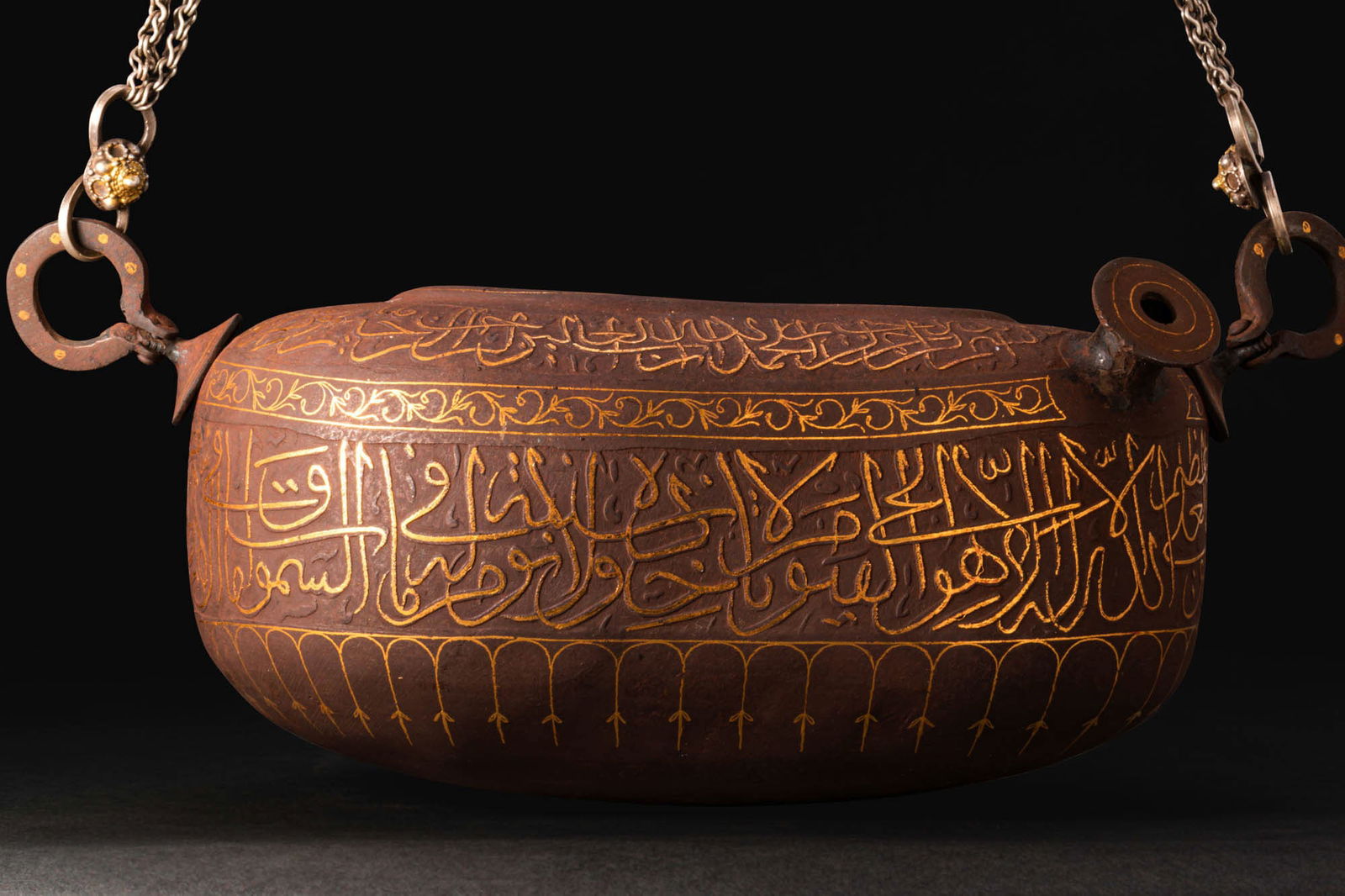 QAJAR INSCRIBED AND INLAID KASHKUL (BEGGAR'S BOWL): Ca. 19th - 20th century AD. A gold-damascened iron kashkul of elongated boat form with a flattened underside and rounded shoulder with a short pouring spout, the ends with pierced lugs linked by suspe