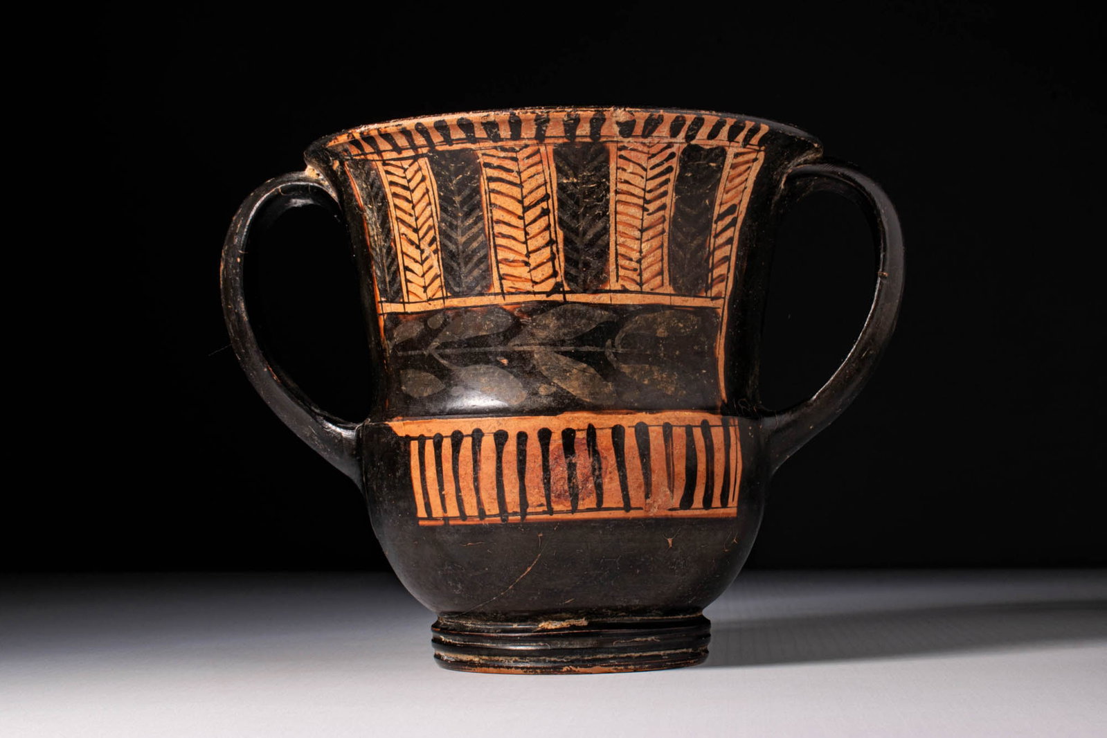 GREEK BLACK GLAZE SESSILE KANTHAROS: Ca. 450 BC. A black glaze sessile kantharos with a rounded lower body, flaring neck and twin horizontal handles. Both sides are decorated with vertical bands of alternating black and reserved chevrons