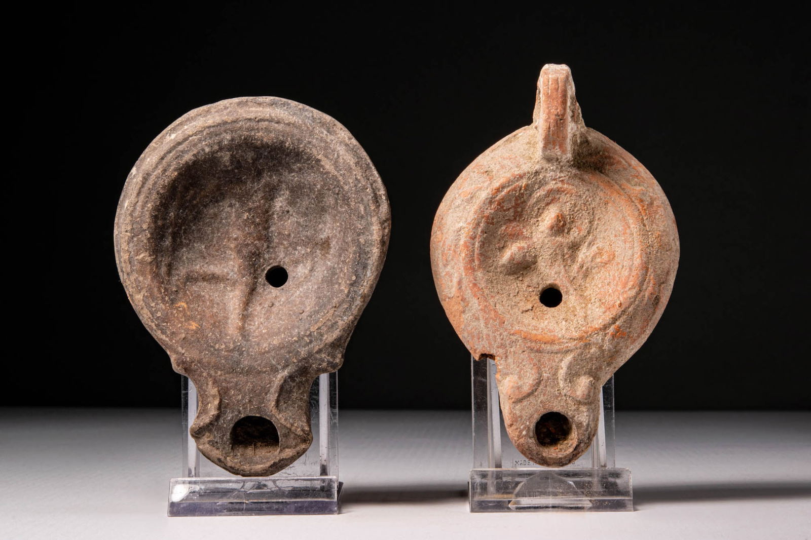 PAIR OF ROMAN TERRACOTTA OIL LAMP: Ca. AD 1 - 200. A pair of terracotta oil lamps. The one on the left is mould-made in dark grey fabric with a simple raised rim and a relief motif on the discus. The lamp on the right, in orange clay,