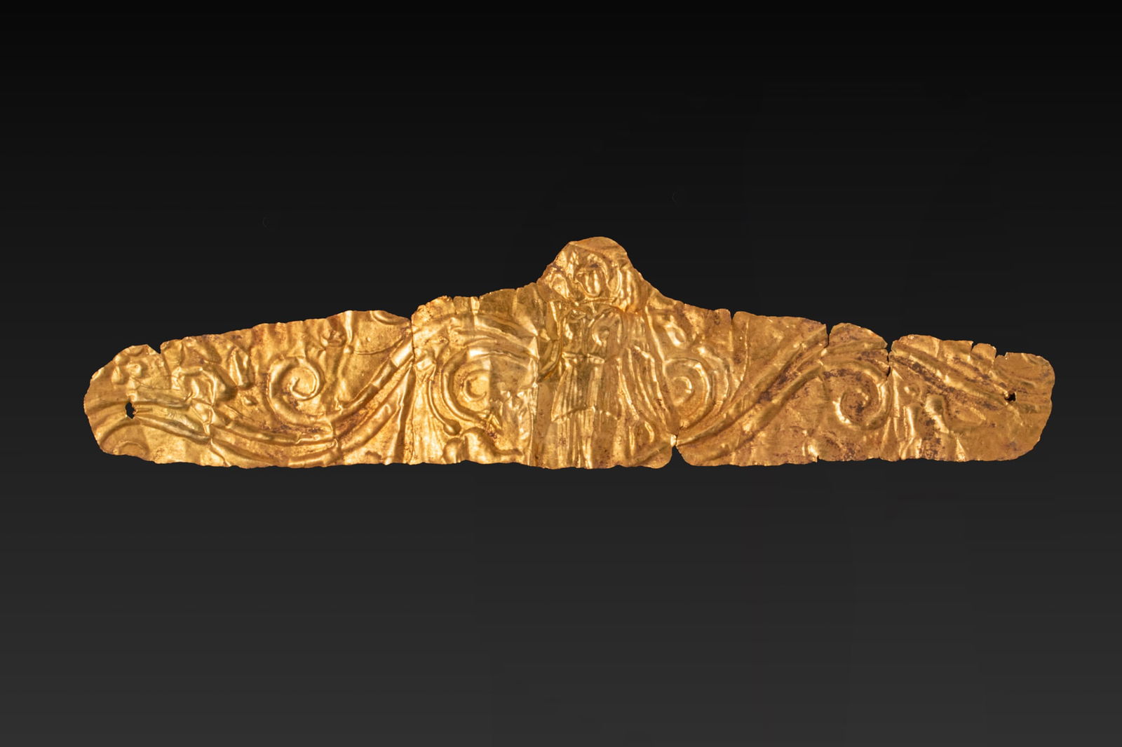 GREEK HIGH KARAT GOLD DIADEM WITH WINGED NIKE AND FLORAL MOTIF: Ca. 350 - 300 BC A gold diadem formed from a thin sheet of gold, with an elongated body and rounded terminals. The band is decorated in repouss? with a central winged Nike standing frontally in a long