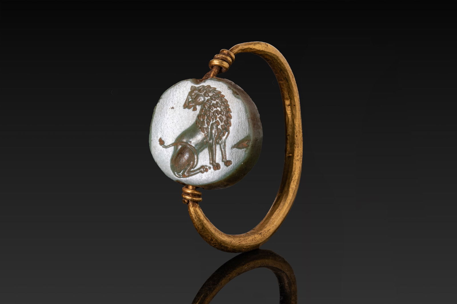GREEK GOLD SWIVEL RING WITH INTAGLIO OF A LION: Ca. 500 BCA gold swivel ring with a slender rounded hoop, the terminals bound with short coils of wire and supporting a transverse bar suspending a circular pale green intaglio. The obverse is engrave