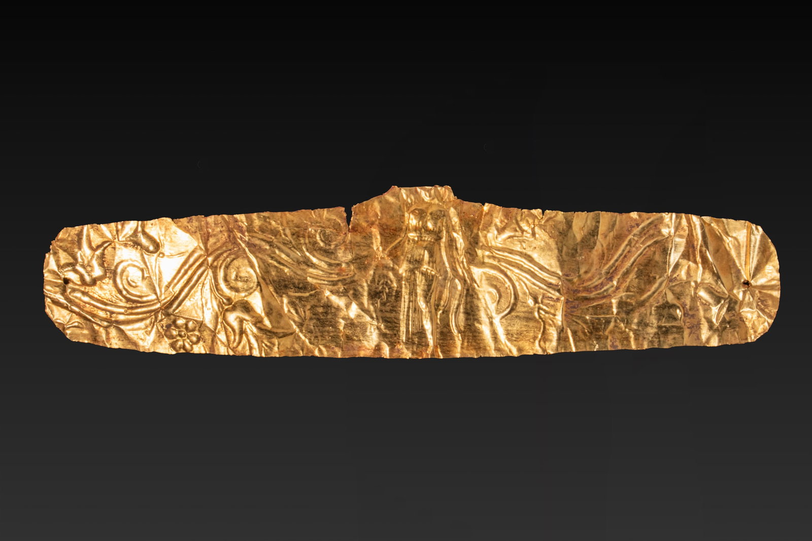 GREEK HIGH KARAT GOLD DIADEM WITH WINGED NIKE AND FLORAL MOTIFS: Ca. 350 - 300 BC A gold diadem formed from a thin sheet of gold, with an elongated body and rounded terminals. The band is decorated in repouss? with a central winged Nike standing frontally in a long