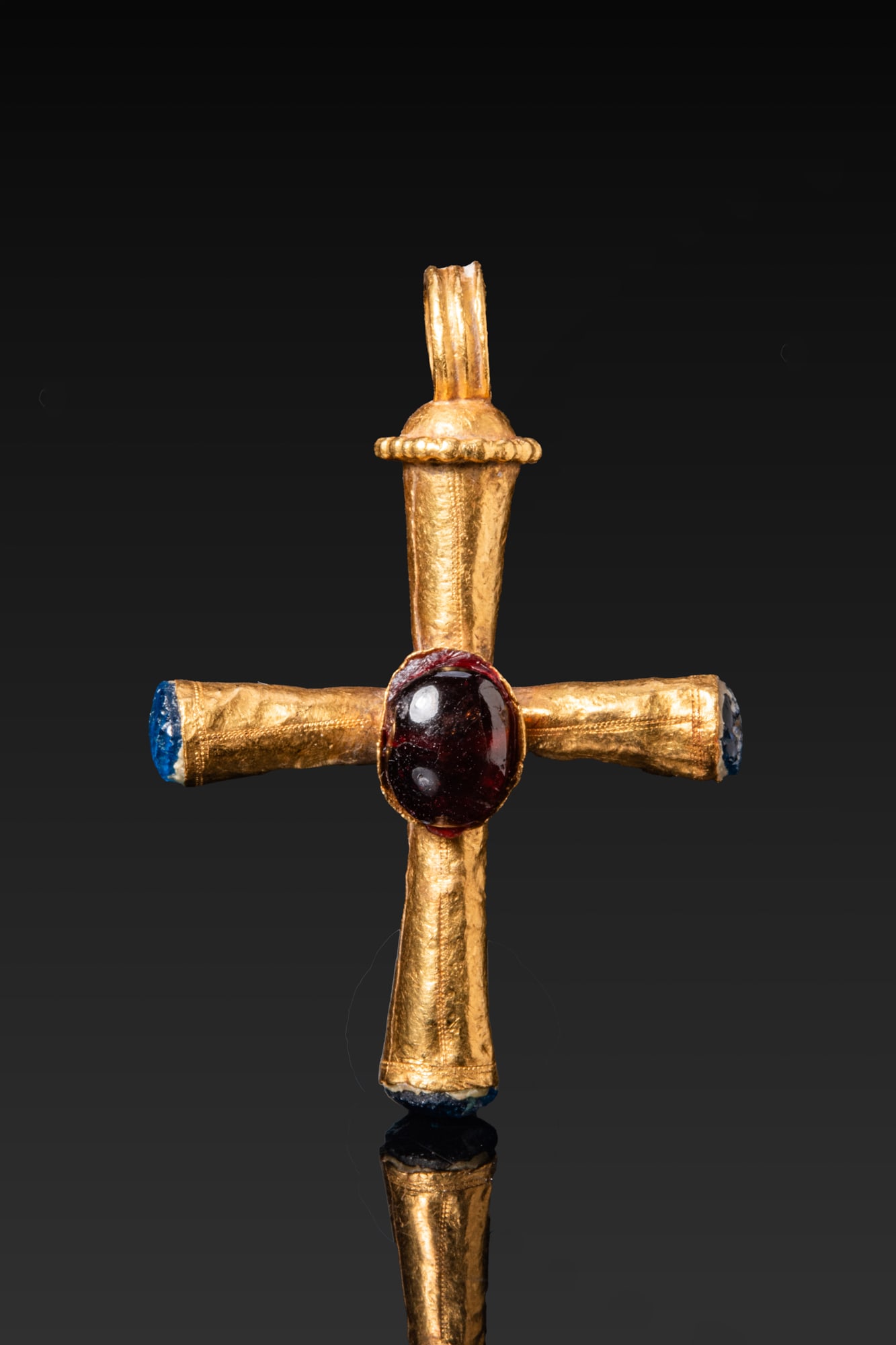 BYZANTINE HIGH KARAT GOLD CROSS PENDANT WITH RED GARNET CABOCHON AND BLUE GLASS FINIALS: Ca. AD 600 A fine gold cross pendant formed from tubular arms slightly flaring outwards. Each arm terminates in a cylindrical socket fitted with a rounded blue glass finial. At the crossing a deep ova