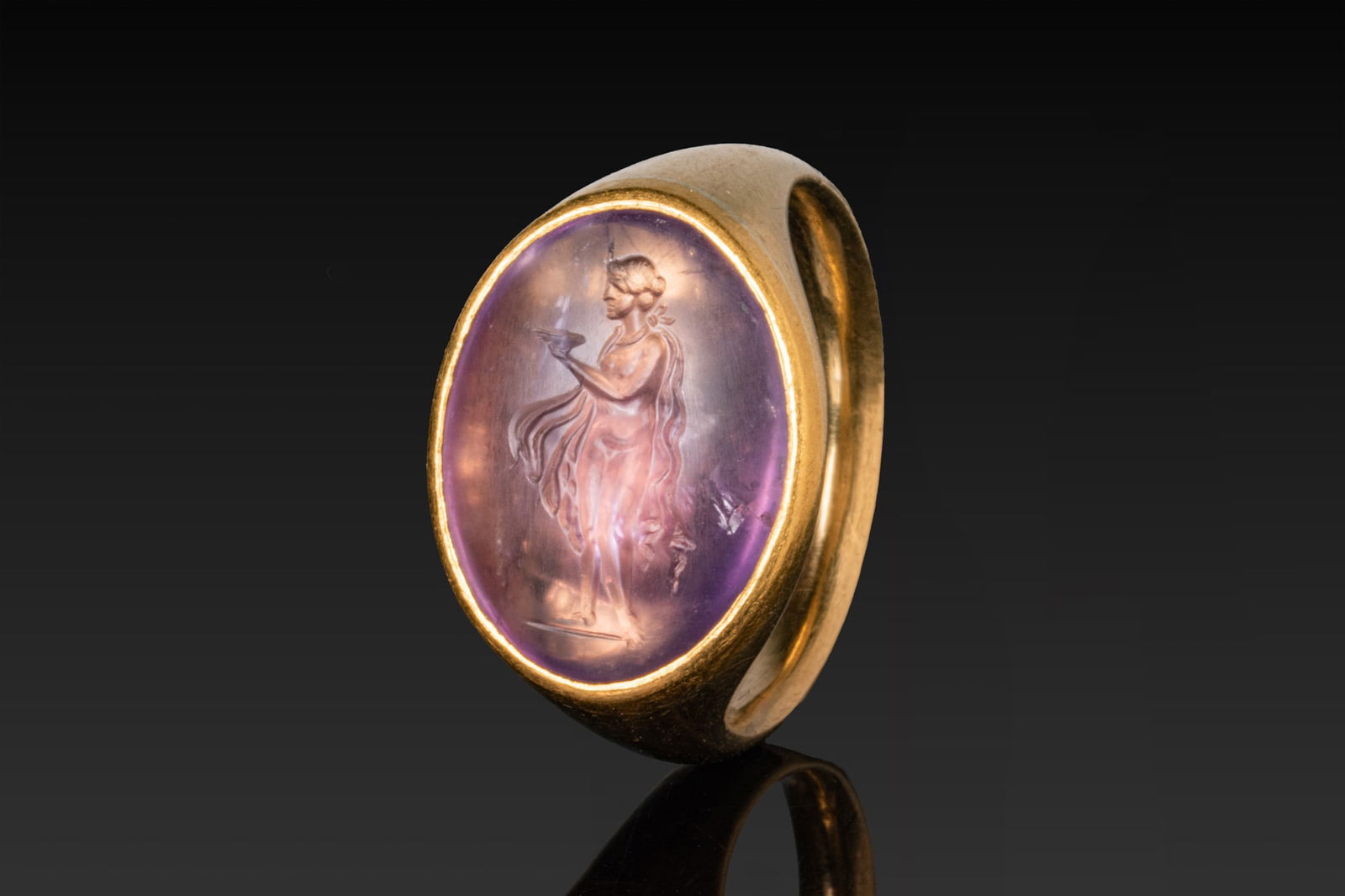 ROMAN AMETHYST INTAGLIO OF A MAENAD MAKING AN OFFERING IN HIGH KARAT GOLD RING: Ca. AD 200 - 250 An amethyst intaglio engraved with a standing maenad in profile, the slender figure nude save for a mantle falling from the shoulders, the left arm extended to hold a shallow offering