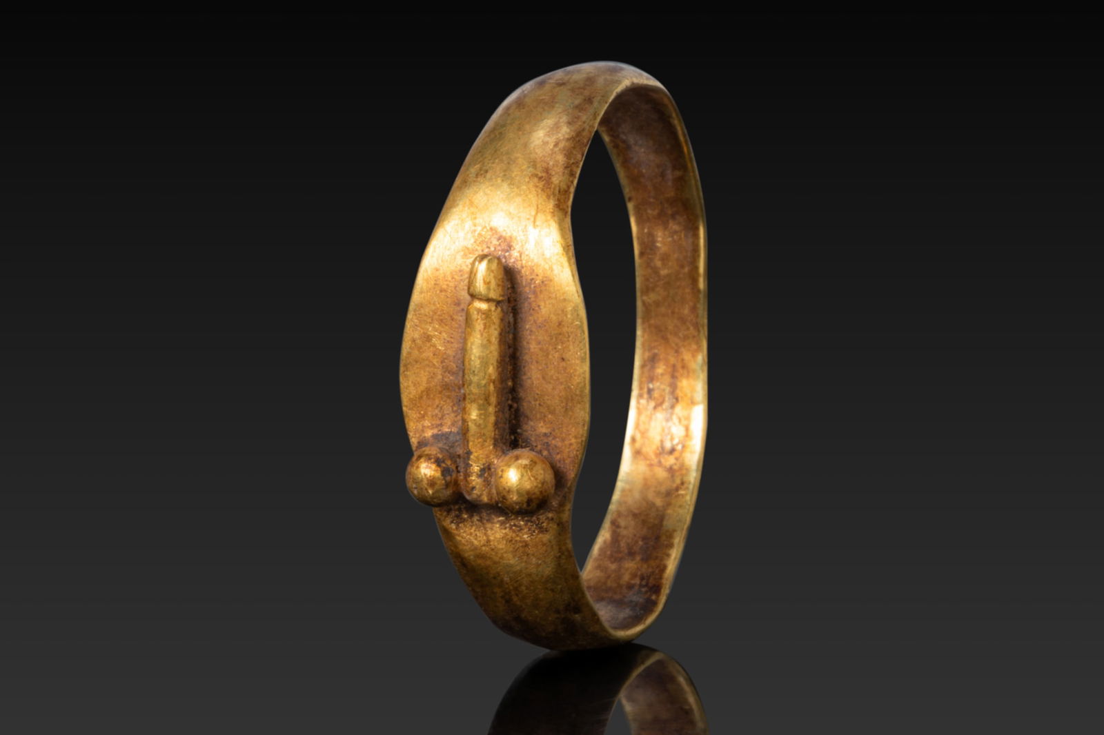 ROMAN HIGH KARAT GOLD RING WITH PHALLUS: Ca. AD 1 - 300 A gold ring with a flat hoop forming an oval bezel decorated in relief with a phallus modelled as a straight shaft with a rounded tip, flanked by a pair of globular testicles. Size: D:2