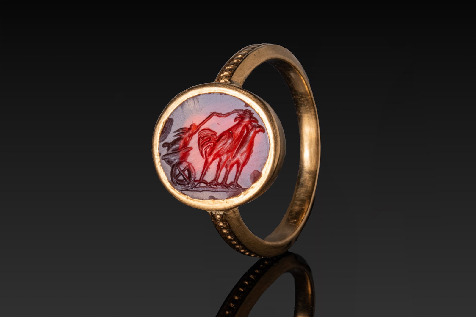 ROMAN CARNELIAN INTAGLIO WITH RABBIT DRIVING A CART DRAWN BY ROOSTERS IN HIGH KARAT GOLD RING: Ca. AD 50 - 200 A carnelian intaglio engraved with a rabbit driving a two-wheeled cart drawn by a pair of roosters, all on a groundline, set within a plain round gold collet of a possibly later gold r