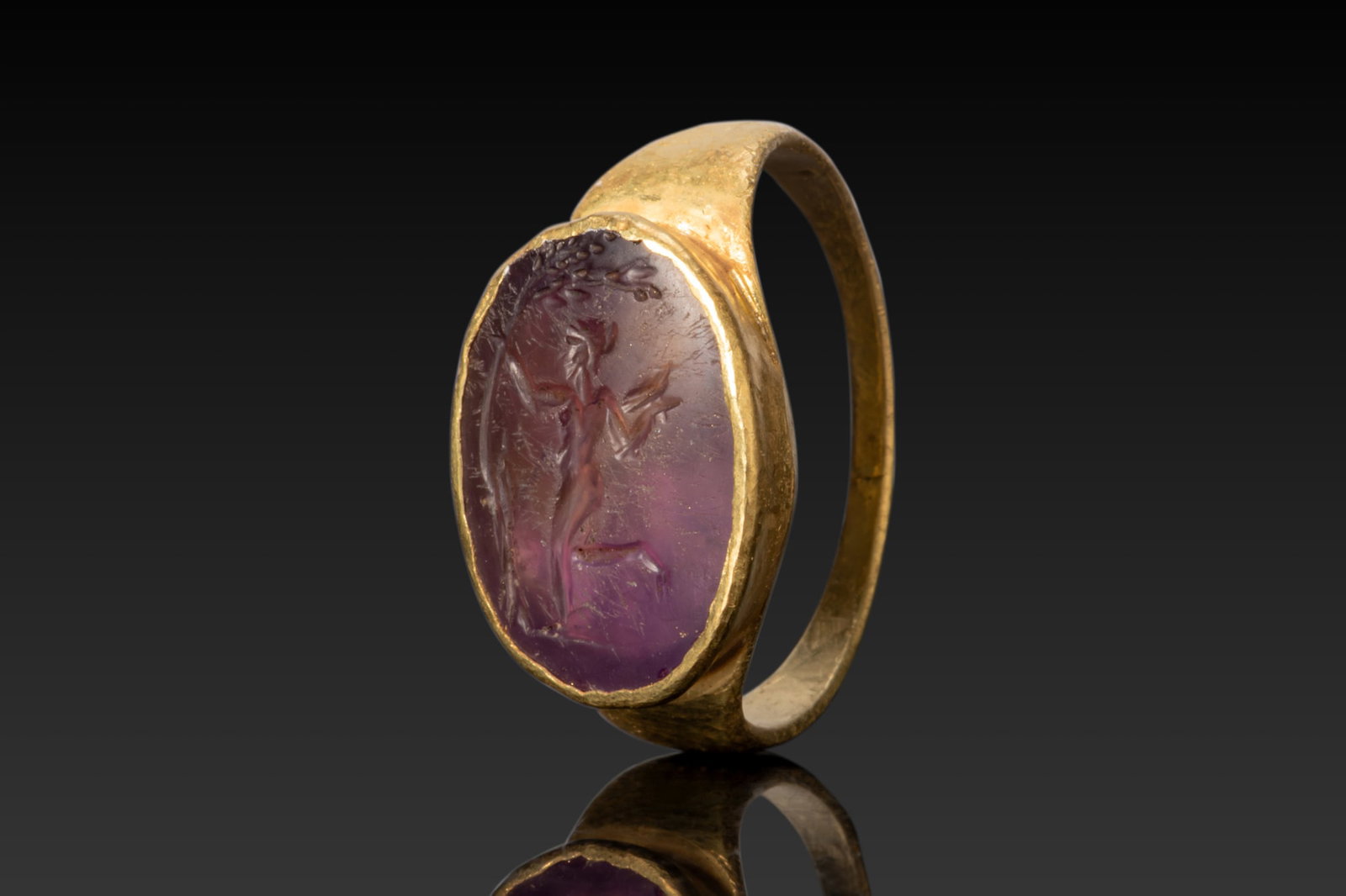 ROMAN HIGH KARAT GOLD RING WITH AMETHYST INTAGLIO OF DANCING MAENAD: Ca. AD 50 - 200 A gold ring with a slender flat-section hoop supporting an oval bezel mount holding an amethyst intaglio in a plain frame. The intaglio is engraved with a dancing maenad in profile on