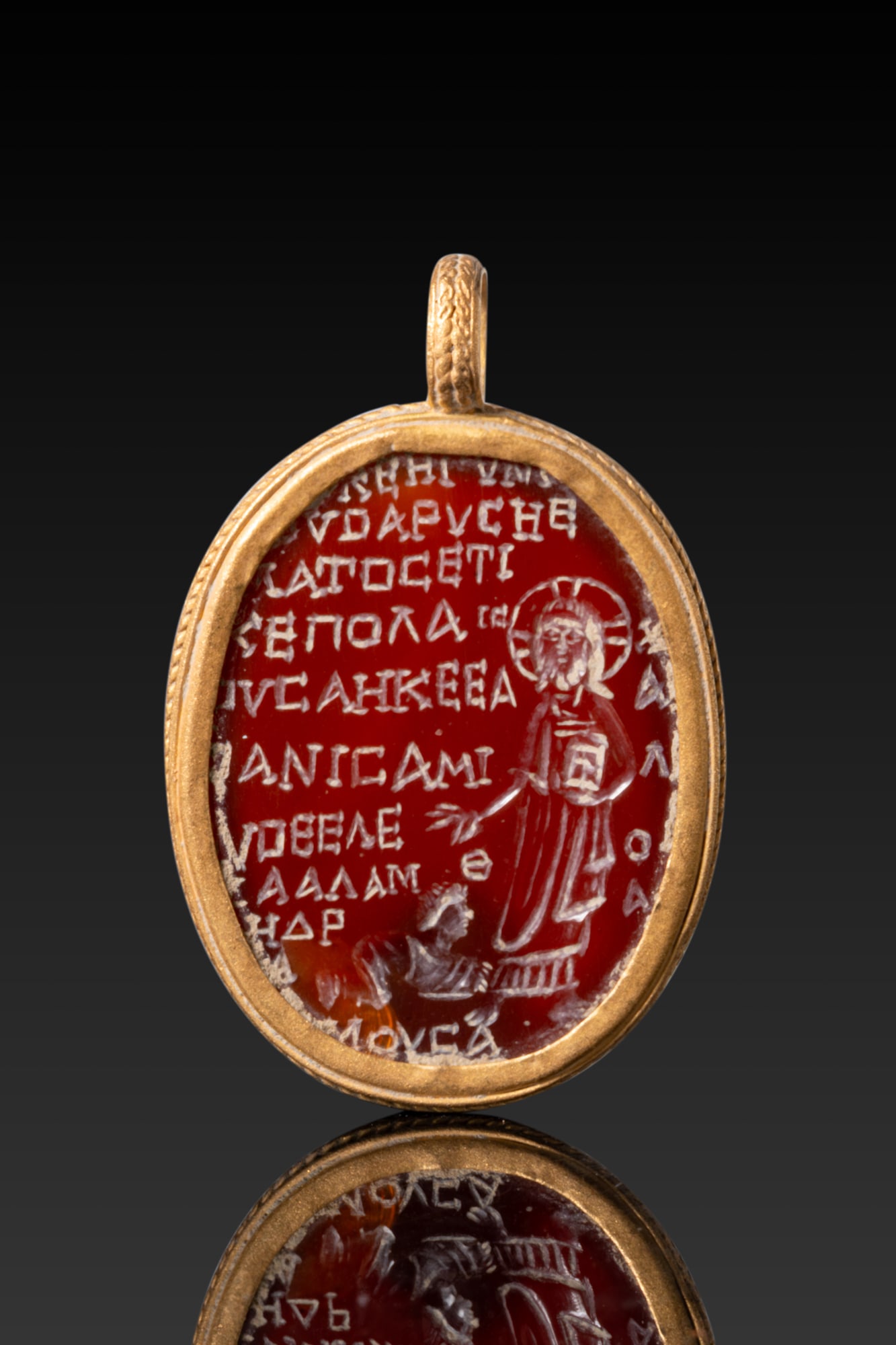BYZANTINE CARNELIAN AMULET WITH CHRIST AND GREEK INSCRIPTION IN GOLD PENDANT: Ca. AD 600A carnelian amulet carved in intaglio with a nimbate Christ standing frontally, holding a book in the left hand and extending the right in blessing over a smaller kneeling figure, the surrou