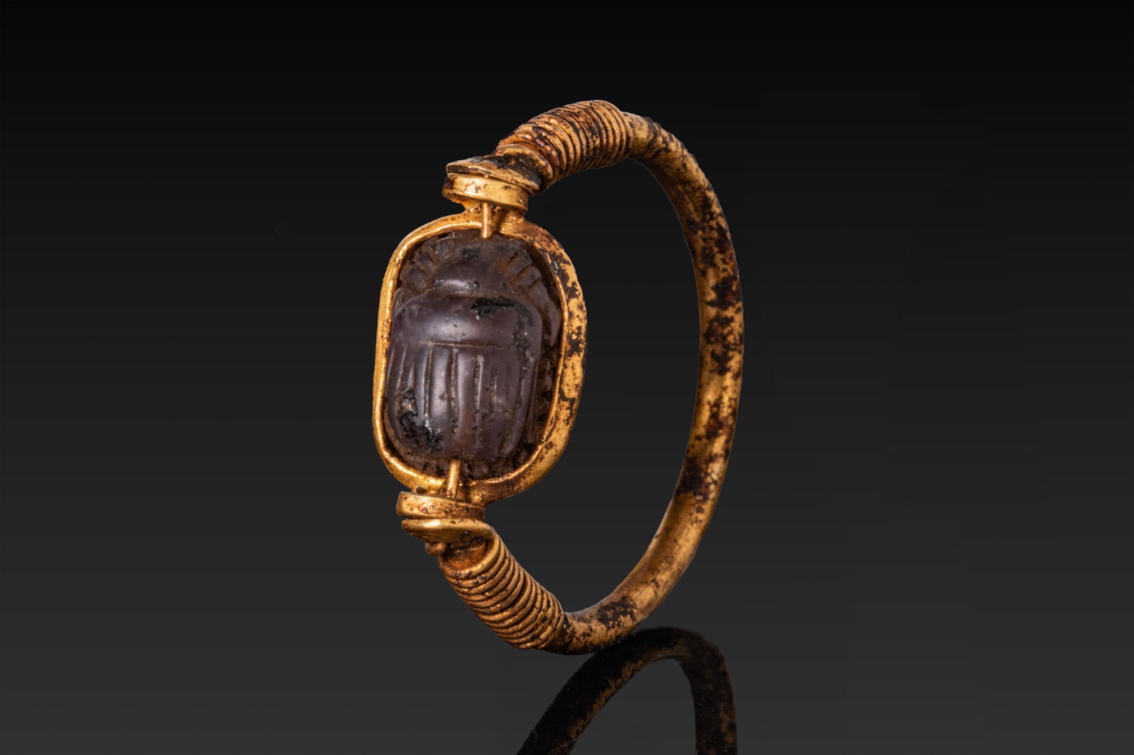 EGYPTIAN GOLD SWIVEL RING WITH INSCRIBED AMETHYST SCARAB: New Kingdom, Ca. 1550 - 1069 BCA gold swivel ring formed from a rounded hoop with its shoulders wrapped in tight spiral wire and terminating in cylindrical collars securing a transverse bar. An amethy