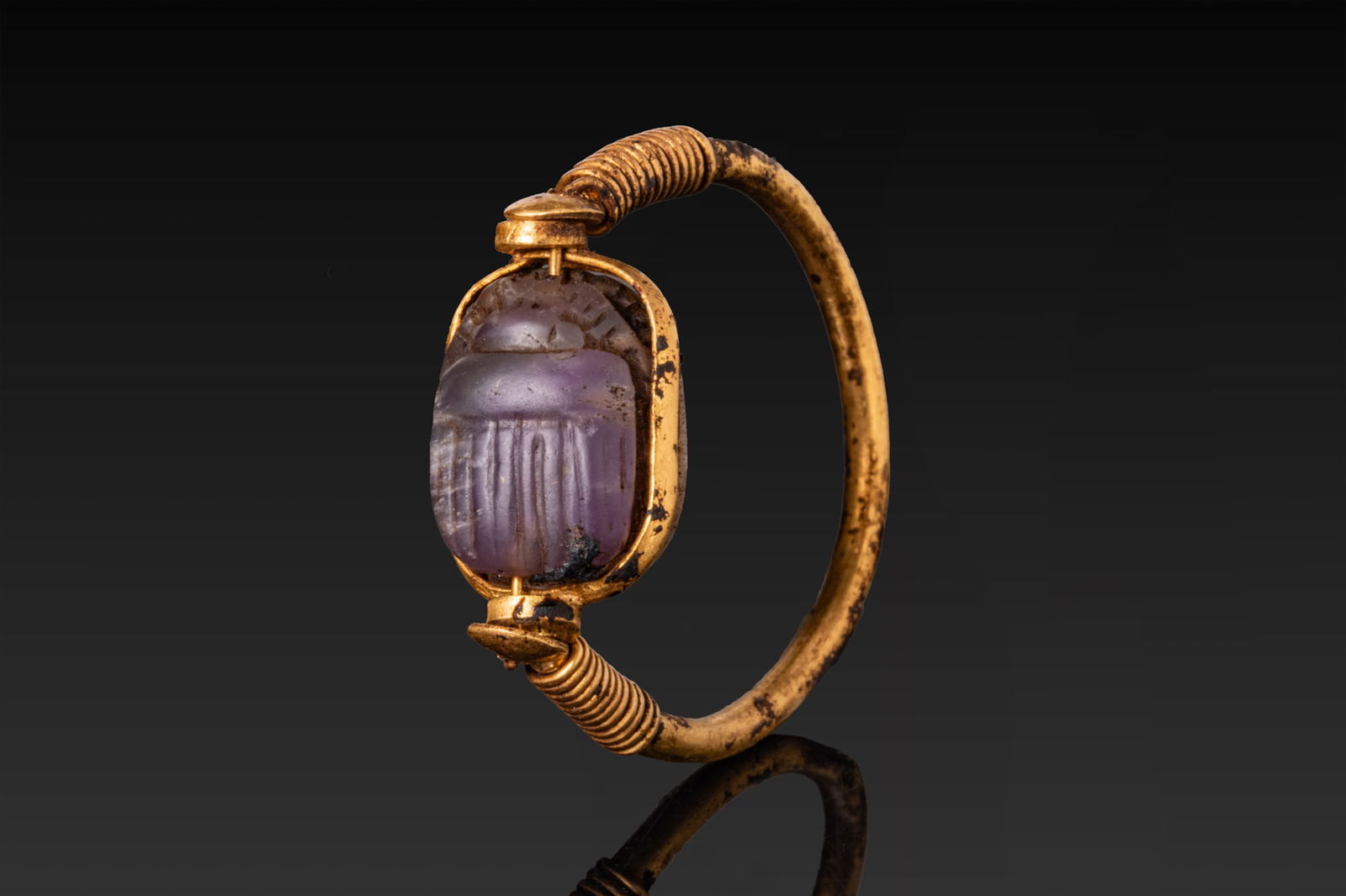 EGYPTIAN GOLD SWIVEL RING WITH INSCRIBED AMETHYST SCARAB: New Kingdom, Ca. 1550 - 1069 BCA gold swivel ring formed from a rounded hoop with its shoulders wrapped in tight spiral wire and terminating in cylindrical collars securing a transverse bar. An amethy