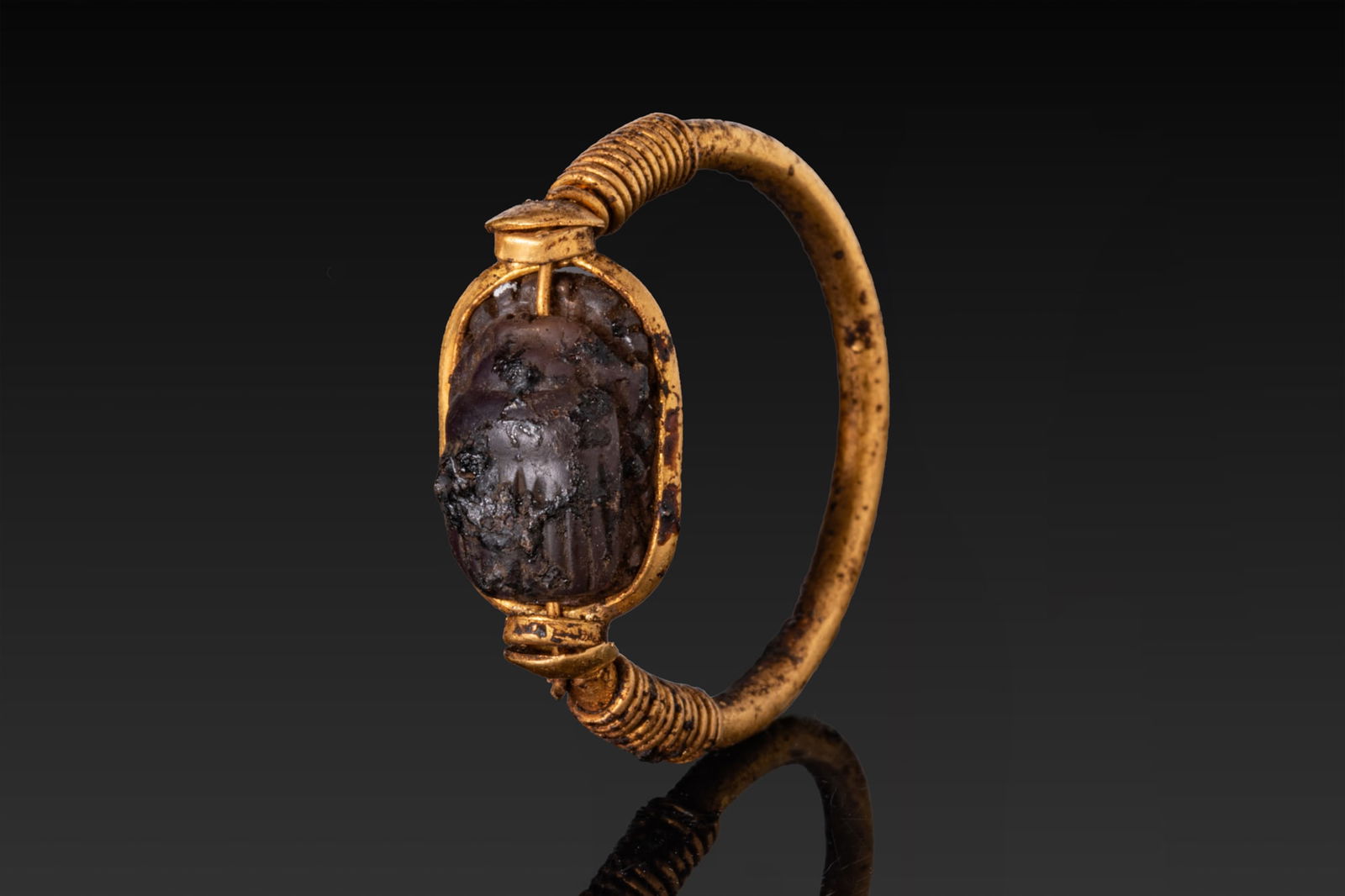 EGYPTIAN GOLD SWIVEL RING WITH INSCRIBED AMETHYST SCARAB: New Kingdom, Ca. 1550 - 1069 BCA gold swivel ring formed from a rounded hoop with its shoulders wrapped in tight spiral wire and terminating in cylindrical collars securing a transverse bar. An amethy