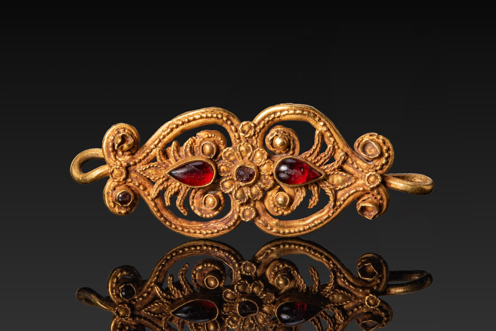 HELLENISTIC HIGH KARAT GOLD PENDANT WITH PALMETTE AND RED GARNET CABOCHONS: Ca. 300 - 100 BC A gold openwork pendant of elongated outline framed by a beaded border. At the centre a rosette in high relief is set with a small round garnet cabochon, flanked on either side by lar