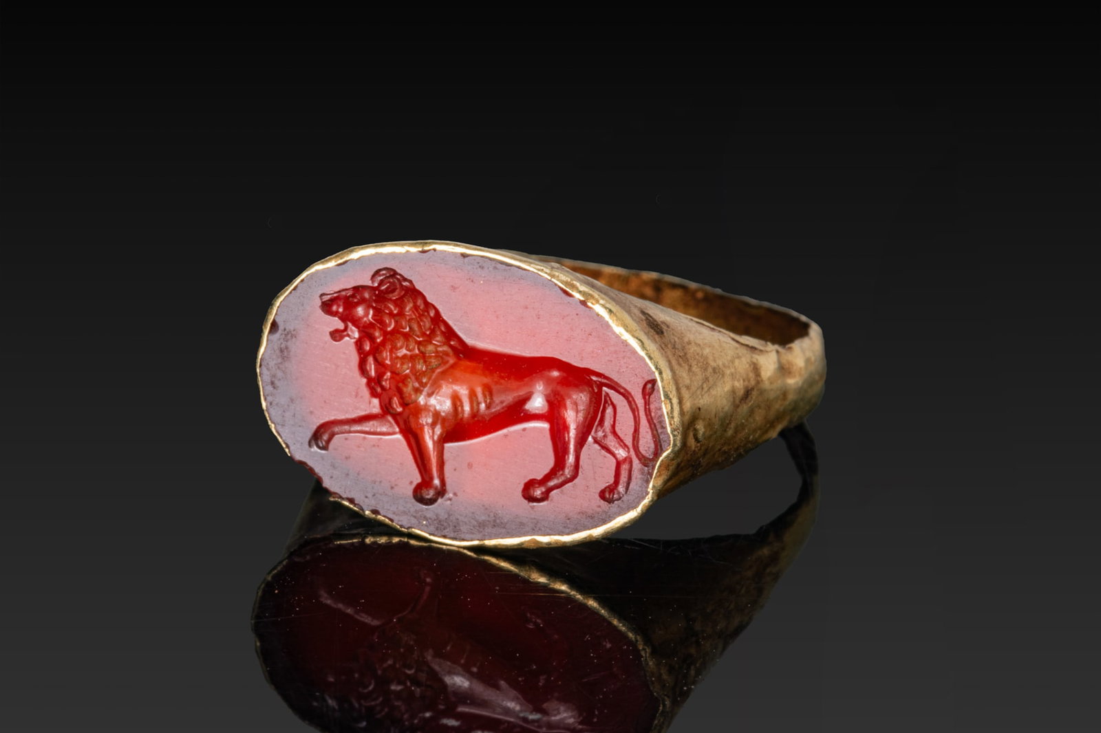 ROMAN GOLD RING WITH CARNELIAN INTAGLIO OF A LION: Ca. 100 BC - AD 100 A gold ring with a flat-section hoop broadening into sloping shoulders that support an oval bezel cell set with a red carnelian intaglio engraved with a lion striding left, the rig