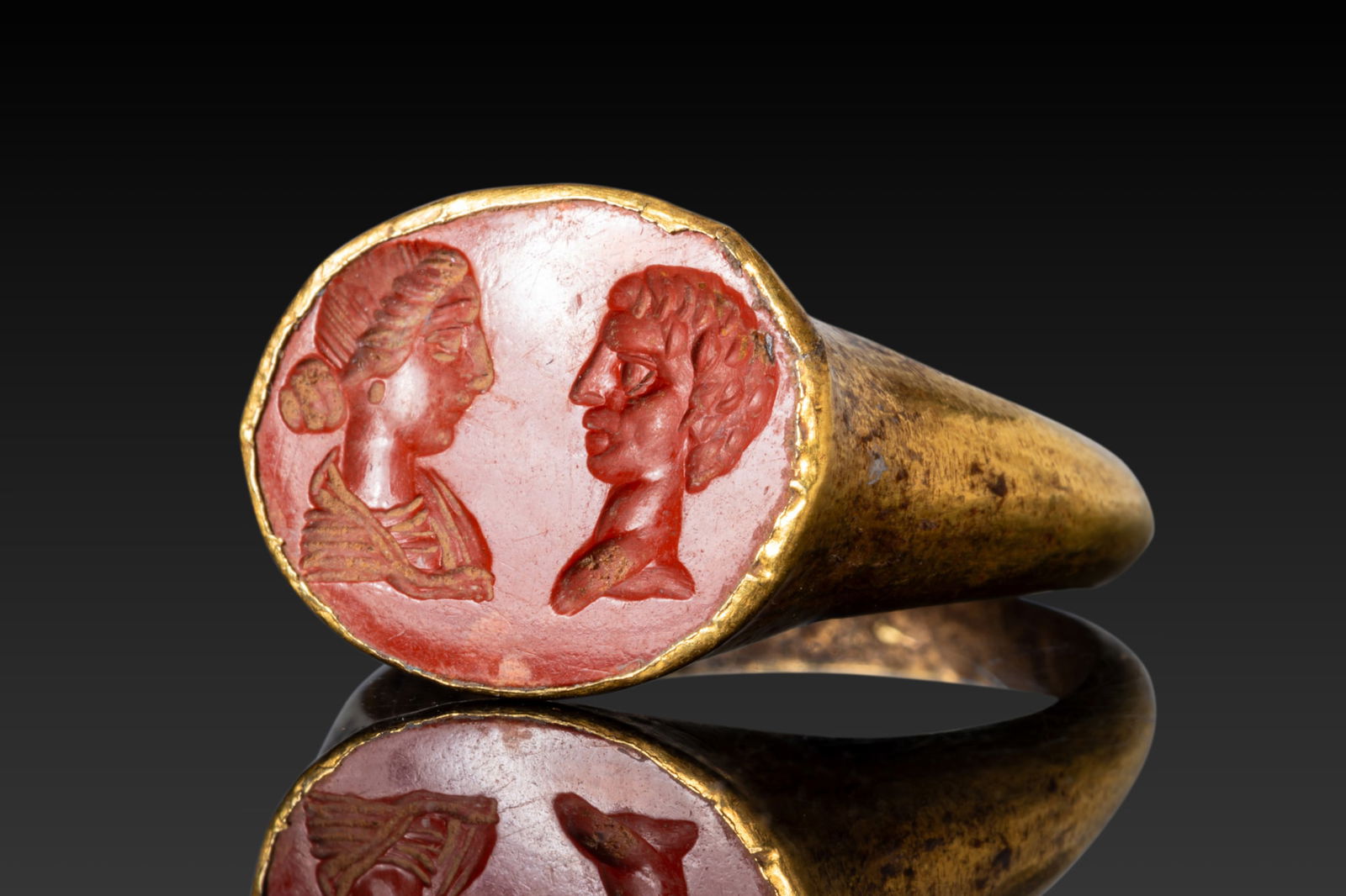 ROMAN HIGH KARAT GOLD RING WITH RED JASPER INTAGLIO DEPICTING A MARRIED COUPLE: Ca. AD 1 - 300 A gold ring with a solid hoop expanding from a narrow rounded shank to broad shoulders and an oval bezel enclosing a red jasper intaglio with confronted busts of a man and woman, the ma