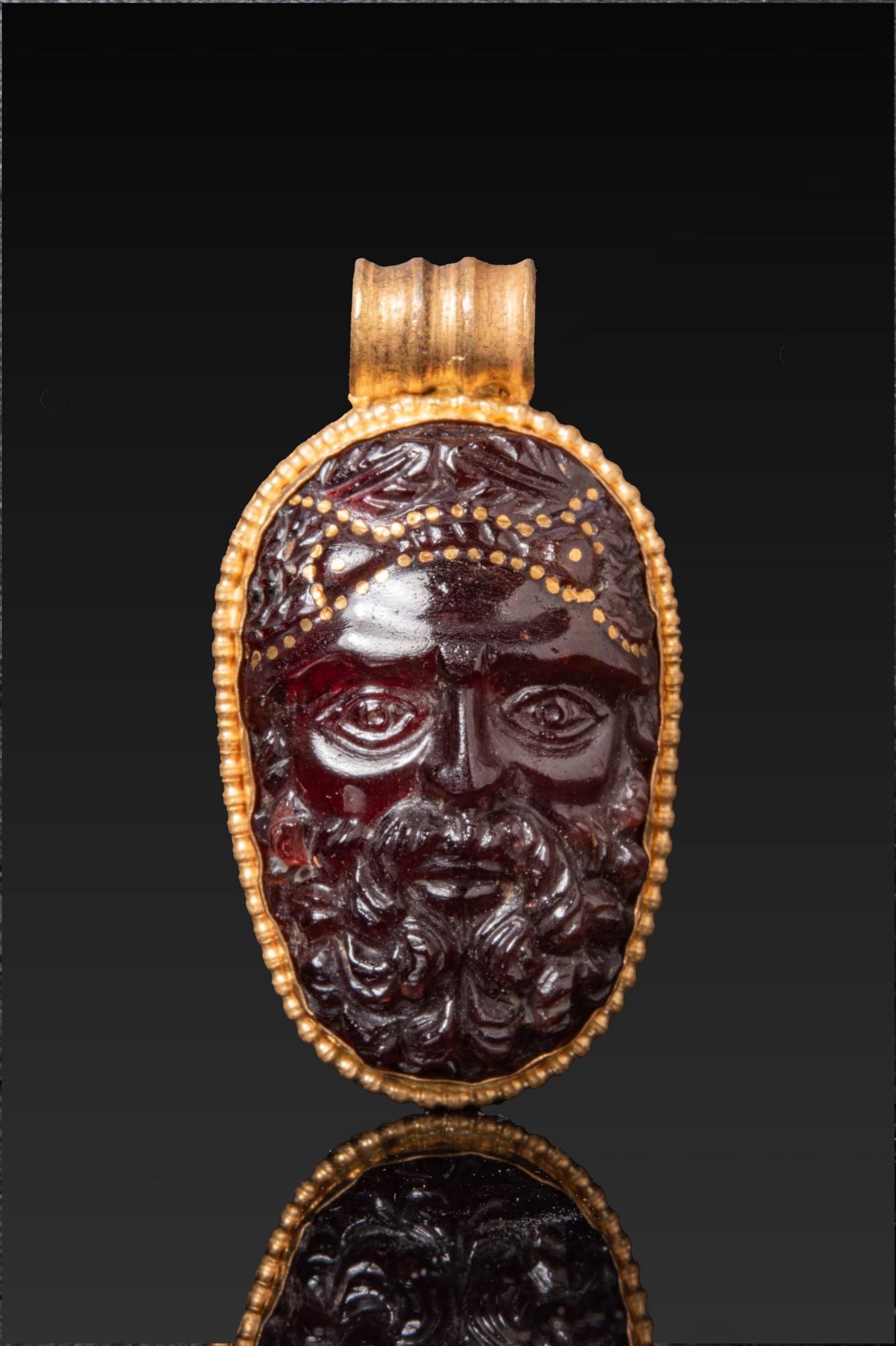 ROMAN RED GARNET CAMEO HEAD OF JUPITER IN HIGH KARAT GOLD PENDANT: Ca. AD 200 - 250 A red garnet cameo carved in high relief with a frontal bearded head of Jupiter, with large eyes, straight nose and full curling beard. The hair is bound with a headband detailed with