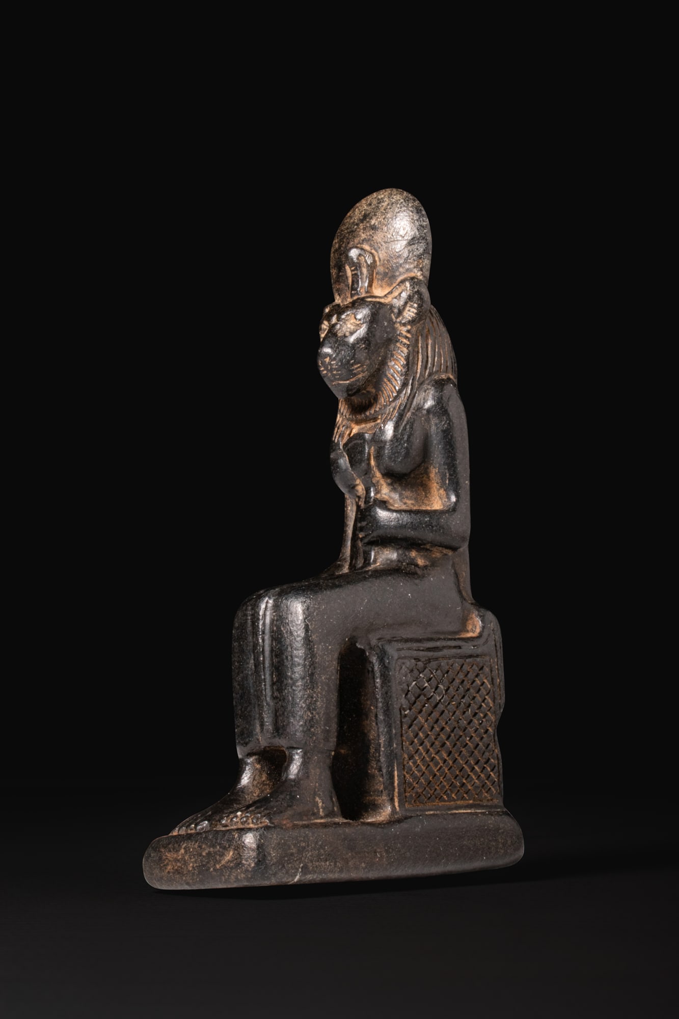 EGYPTIAN BLACK STONE SEATED FIGURE OF SEKHMET: New Kingdom, Ca. 1550 - 1069 BCA black stone figure of Sekhmet seated on a block throne with a low back pillar and cross-hatched side panels, her legs together and feet resting on an integral base.