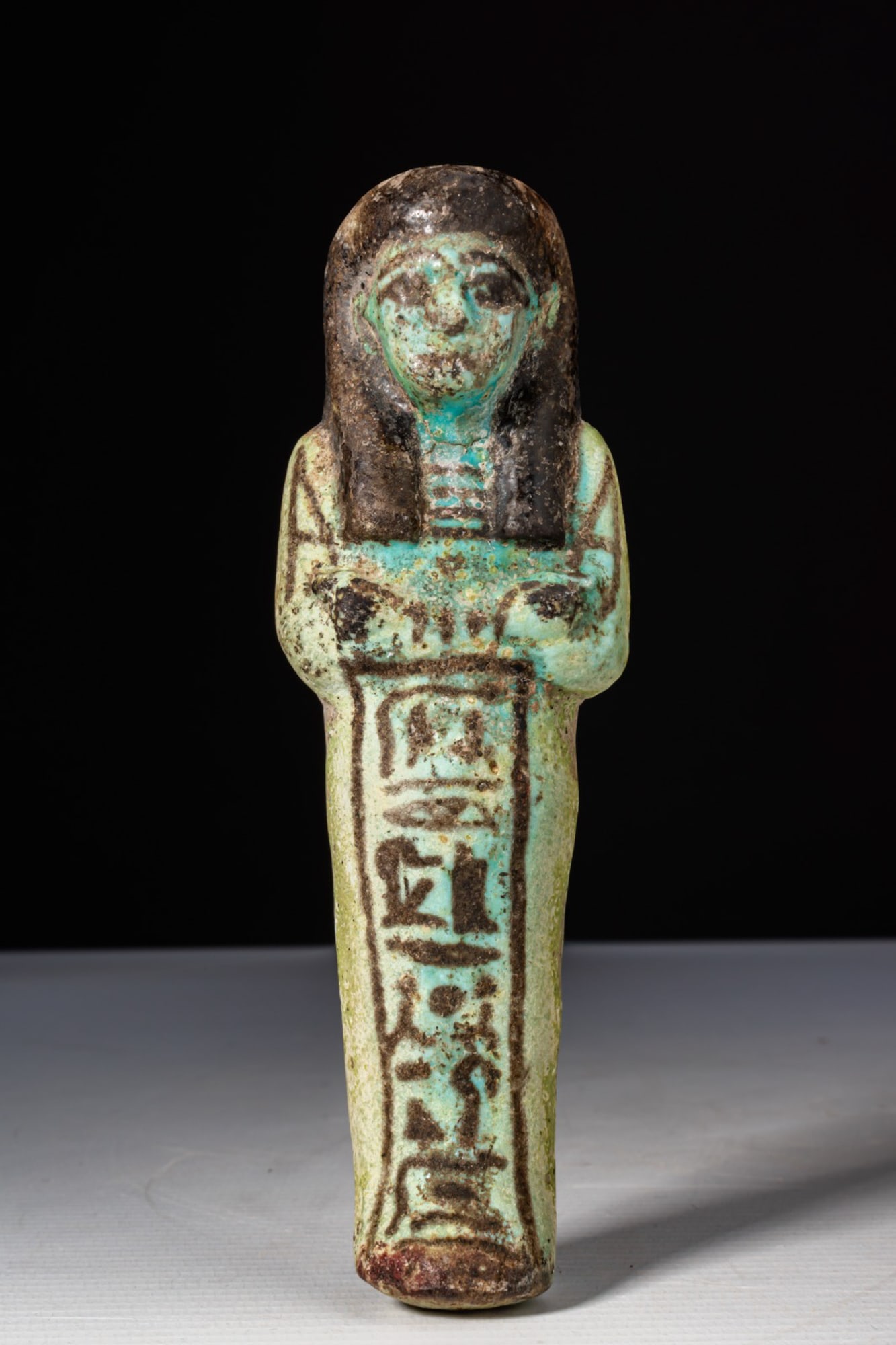 EGYPTIAN FAIENCE USHABTI WITH BLACK DETAILS: Third Intermediate Period, Ca. 1069 - 664 BC A faience ushabti with a black-painted tripartite wig and facial features, shown mummiform with the arms crossed over the chest. The hands are modelled in