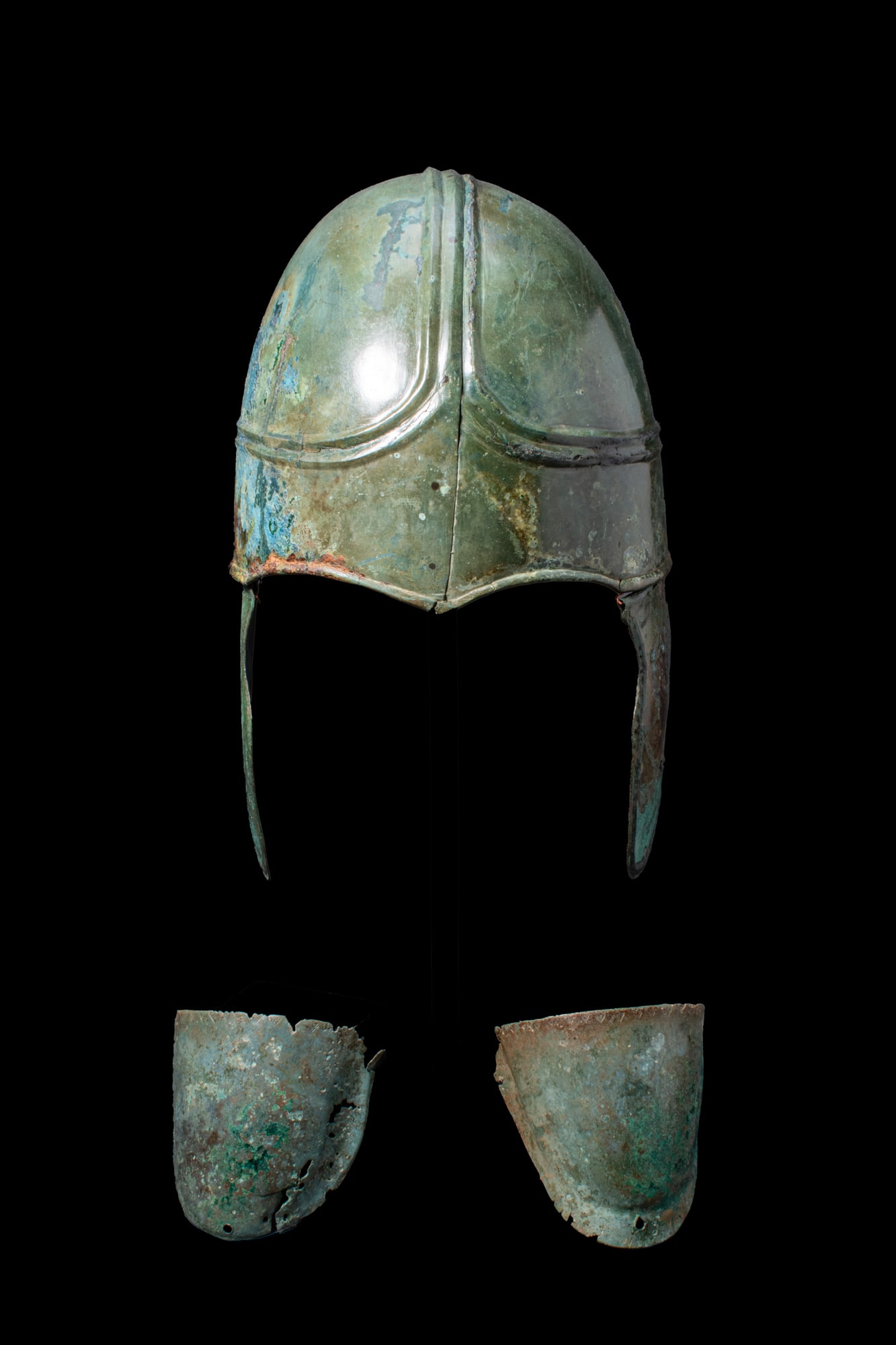 GREEK BRONZE CHALCIDIAN HELMET AND PAIR OF BRONZE SHOULDER PADS: Ca. 250 - 200 BCA bronze Chalcidian helmet with a rounded skull formed in two halves joined along a central ridge. The crown with a raised moulding that bifurcates above the brow and continues around